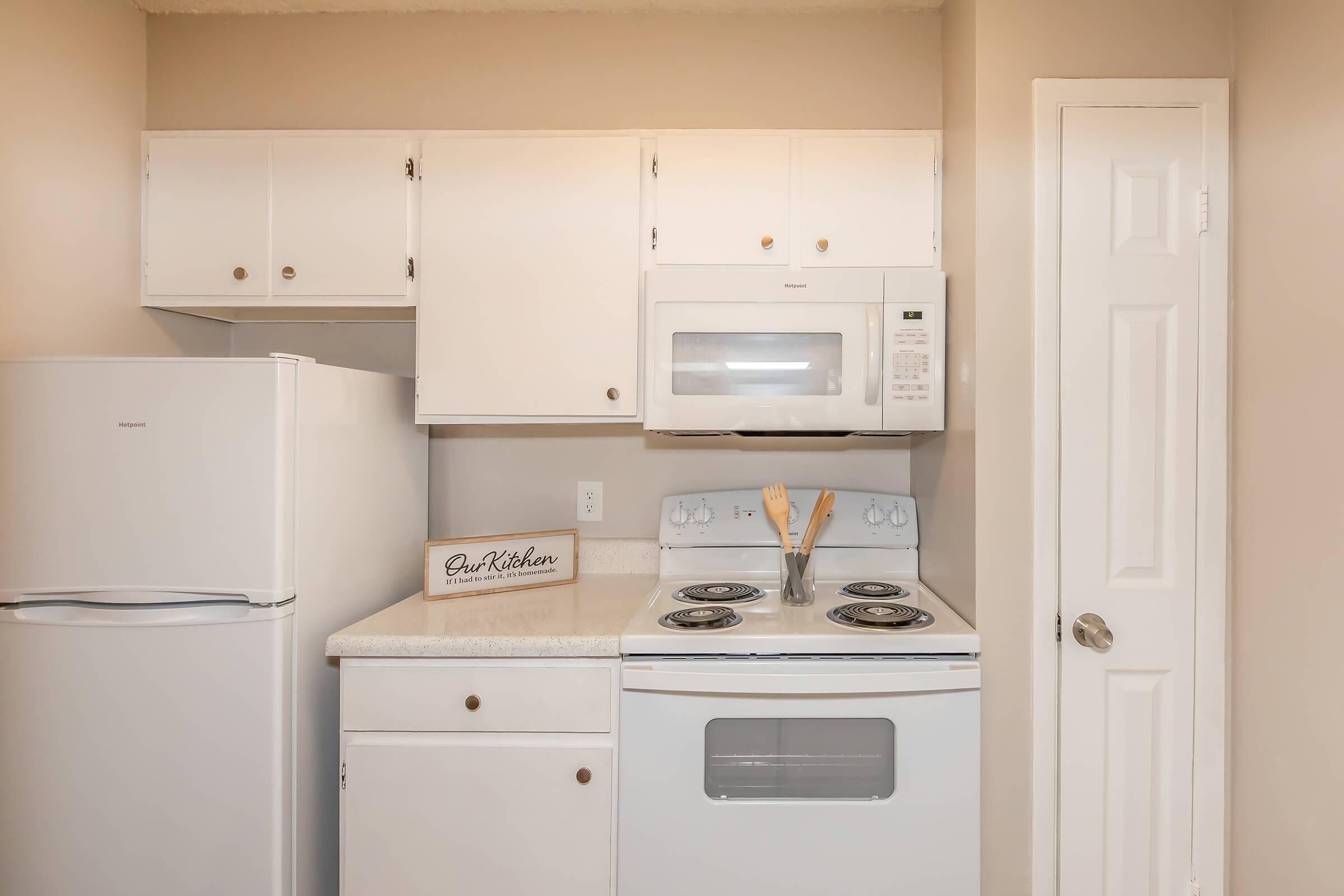 A clean, modern kitchen with white cabinets and appliances. The setup includes a refrigerator, microwave, and stove with an oven, featuring a countertop. A wooden sign reading "Our Kitchen" is placed on the counter, creating a welcoming atmosphere. The walls are painted in a light color, enhancing the brightness of the space.
