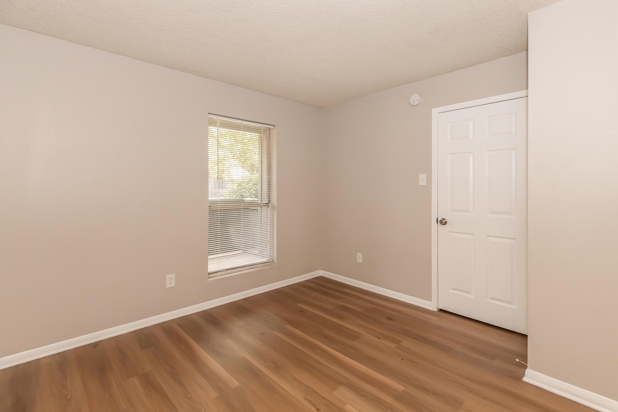 A simple, empty room with light gray walls, a window letting in natural light, and a wooden floor. There is a closed door on the right side of the image, and no furniture or decor present, creating a clean and spacious atmosphere.
