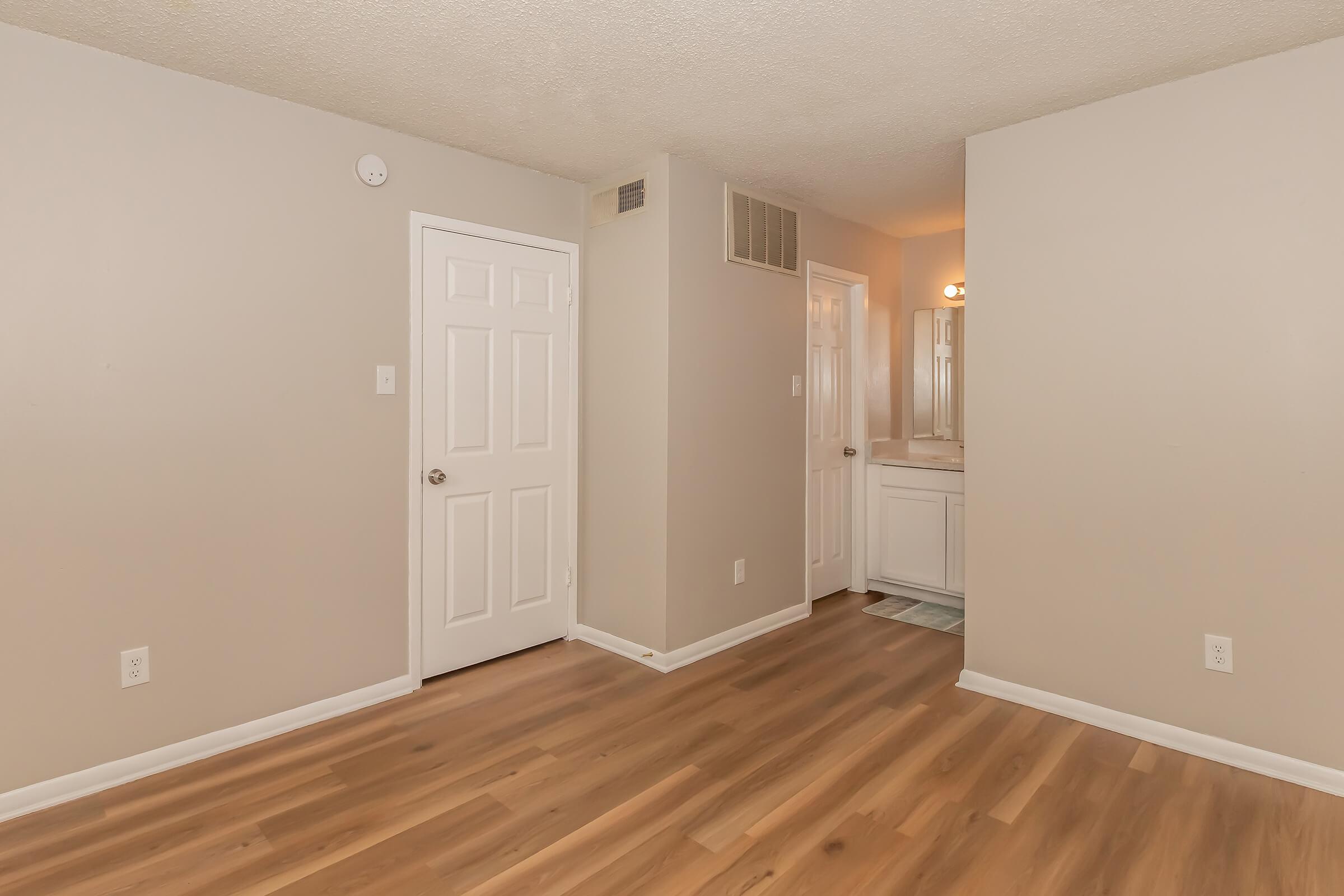 An empty room with light brown wooden flooring, beige walls, and a white door. There is a bathroom door visible on the right side, and the space is well-lit, creating a clean and modern atmosphere.