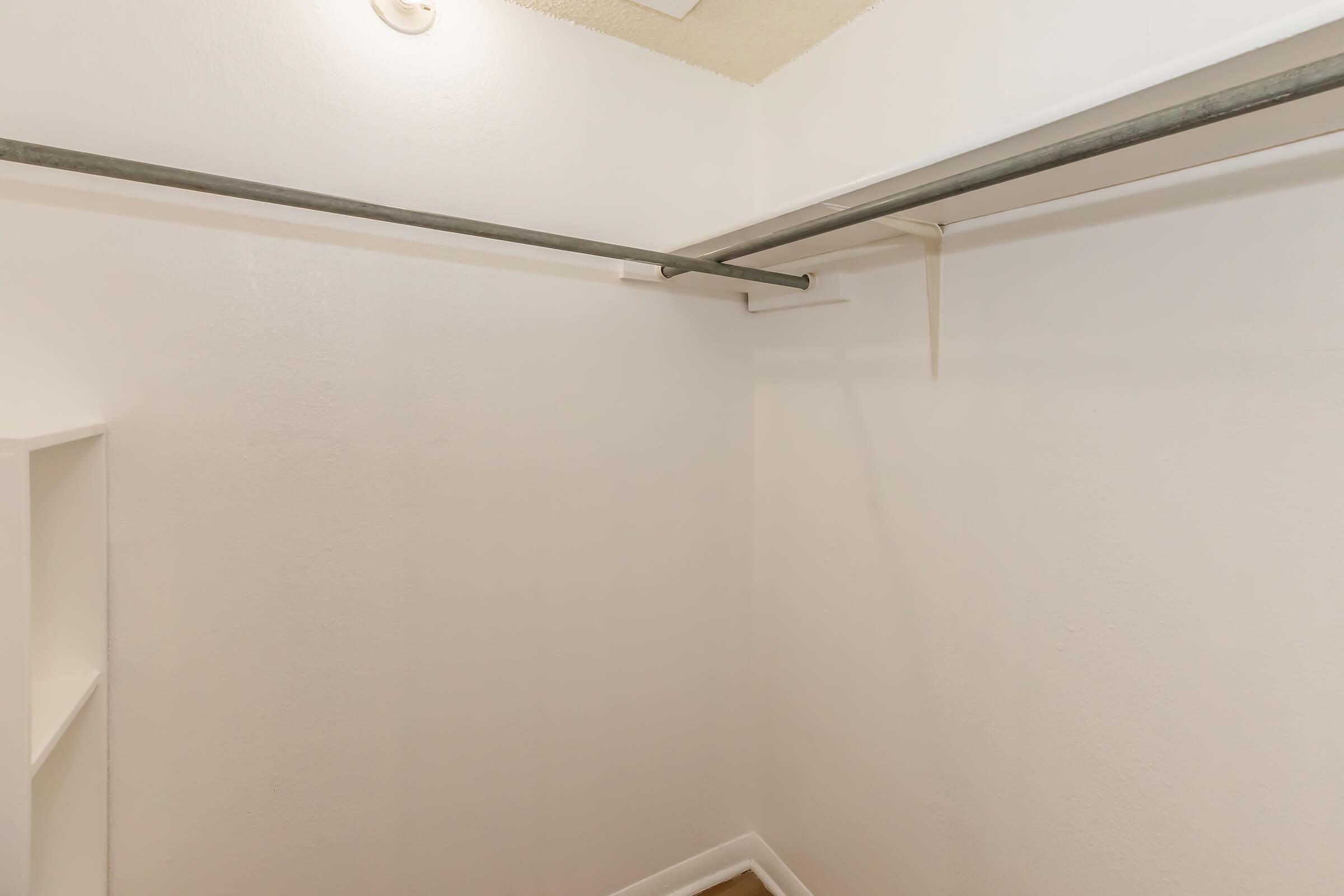 A simple, empty closet with white walls and a metal hanging rod. There is a small shelf on the left side, and the floor is bare. The lighting is soft and neutral, creating a clean and spacious appearance.