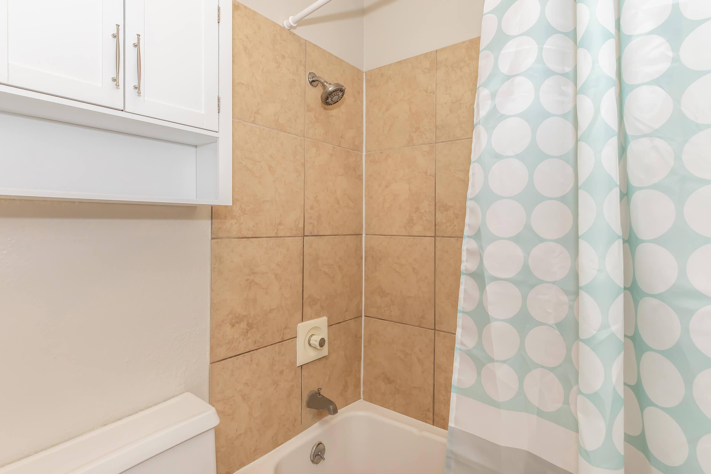 A bathroom with a bathtub, a showerhead, and a light blue shower curtain featuring white polka dots. The walls are tiled with a beige finish, and there's a storage cabinet above. The tub has a faucet, and the overall space appears clean and well-maintained.