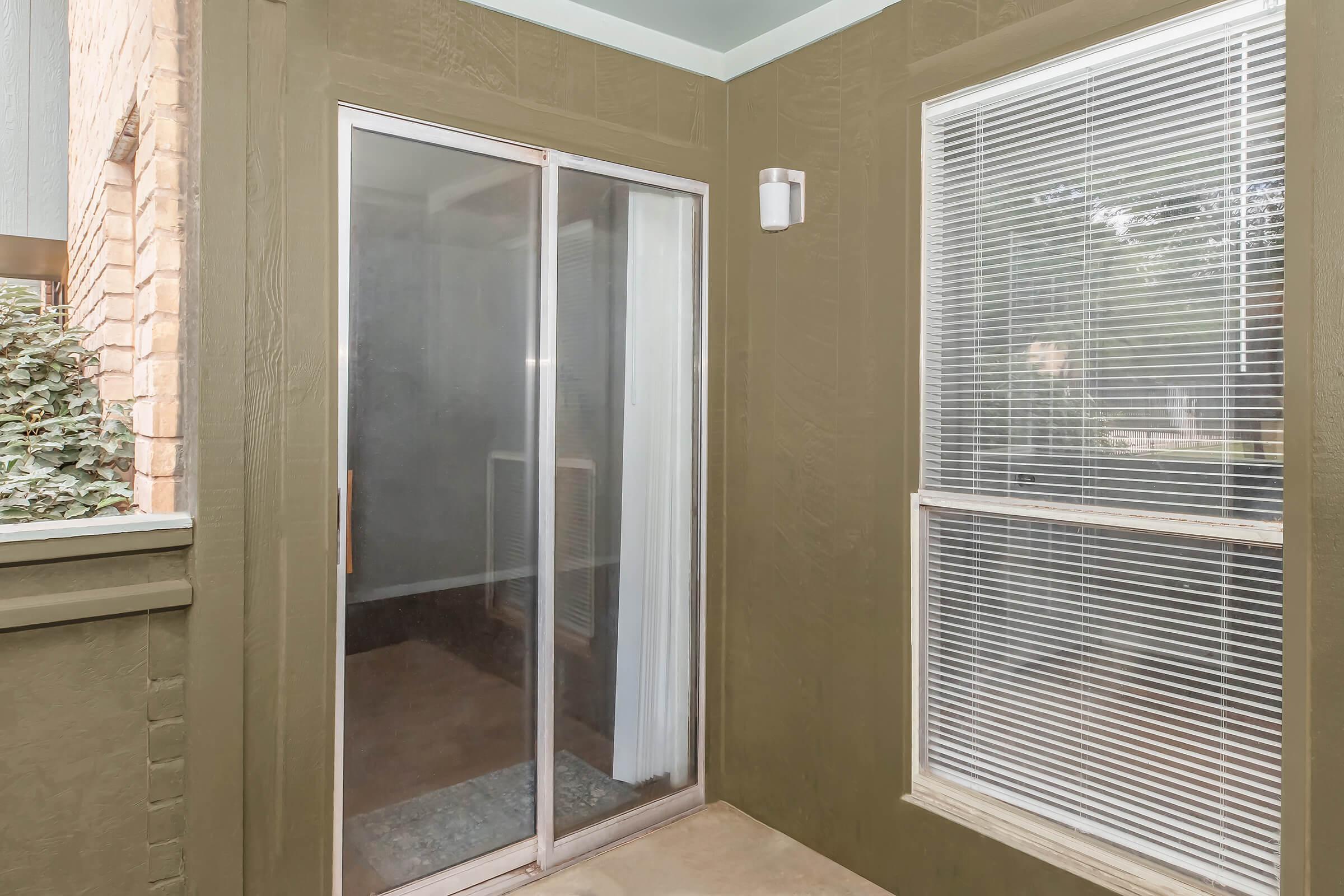 A corner balcony or patio area featuring a sliding glass door and two windows with blinds. The walls are painted olive green, and there's a light fixture on one wall. The flooring appears to be concrete, and a small portion of greenery is visible outside. The space is bright and airy.