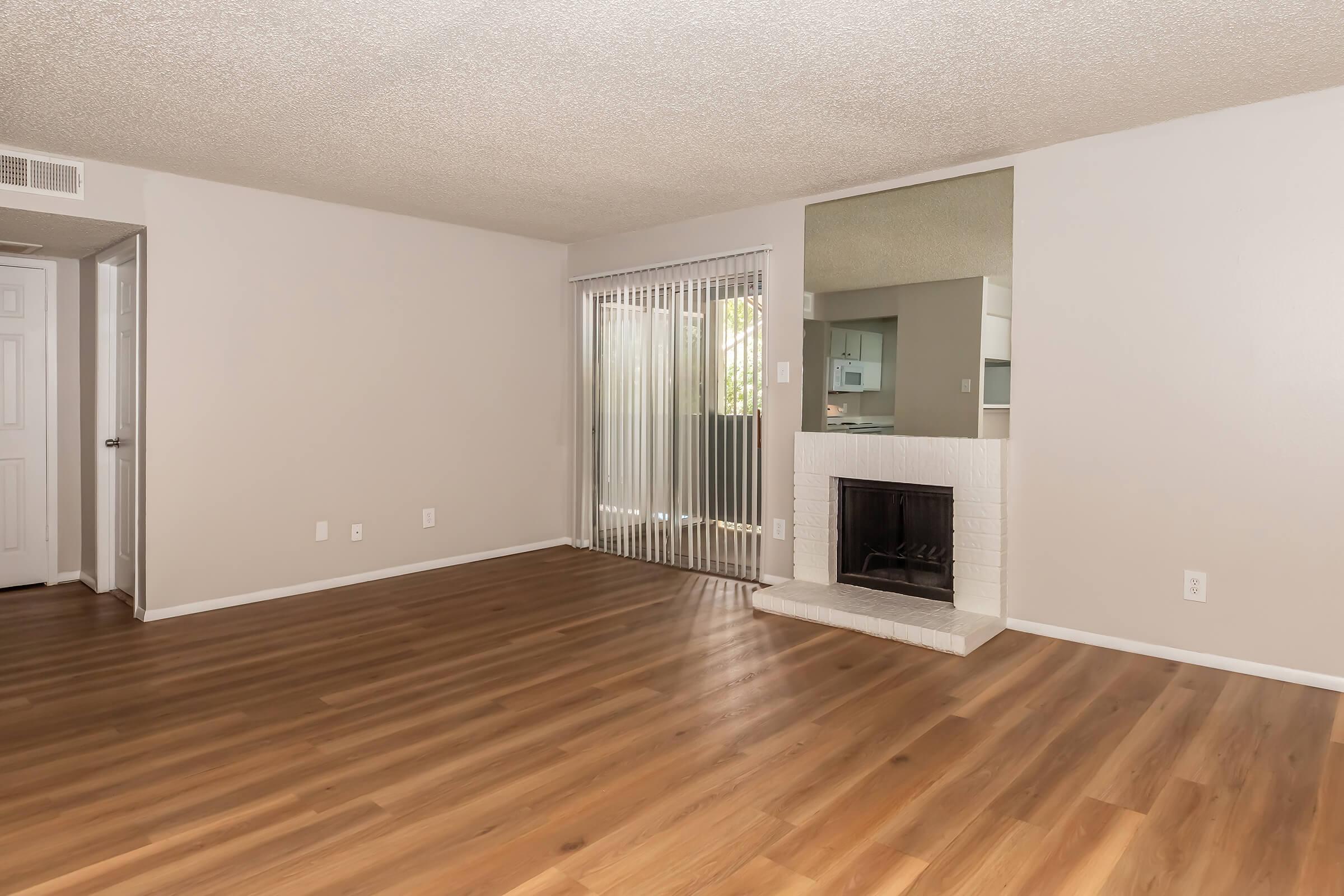 Empty living room with light-colored walls and a large window covered by vertical blinds. The floor is made of light-colored wood. A white brick fireplace is located on the right side of the room, and there is a mirror on the wall above it, reflecting the space. The room appears bright and spacious.