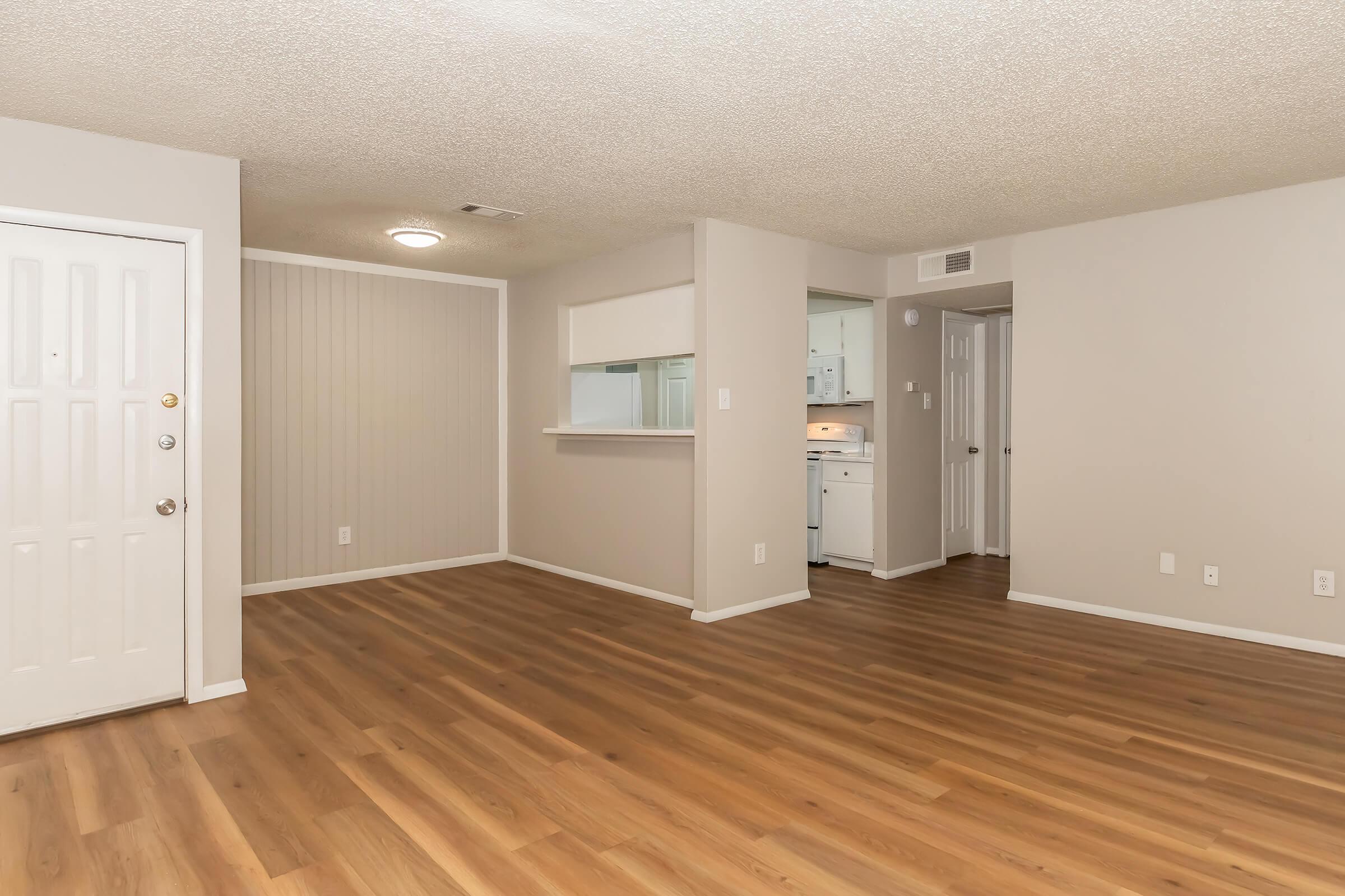 A spacious living room with light-colored walls and a textured ceiling, featuring wooden flooring. A front door is visible on the left, and an open kitchen area is seen in the background through an archway. The room is well-lit with natural light and minimal furniture, providing a clean and open feel.