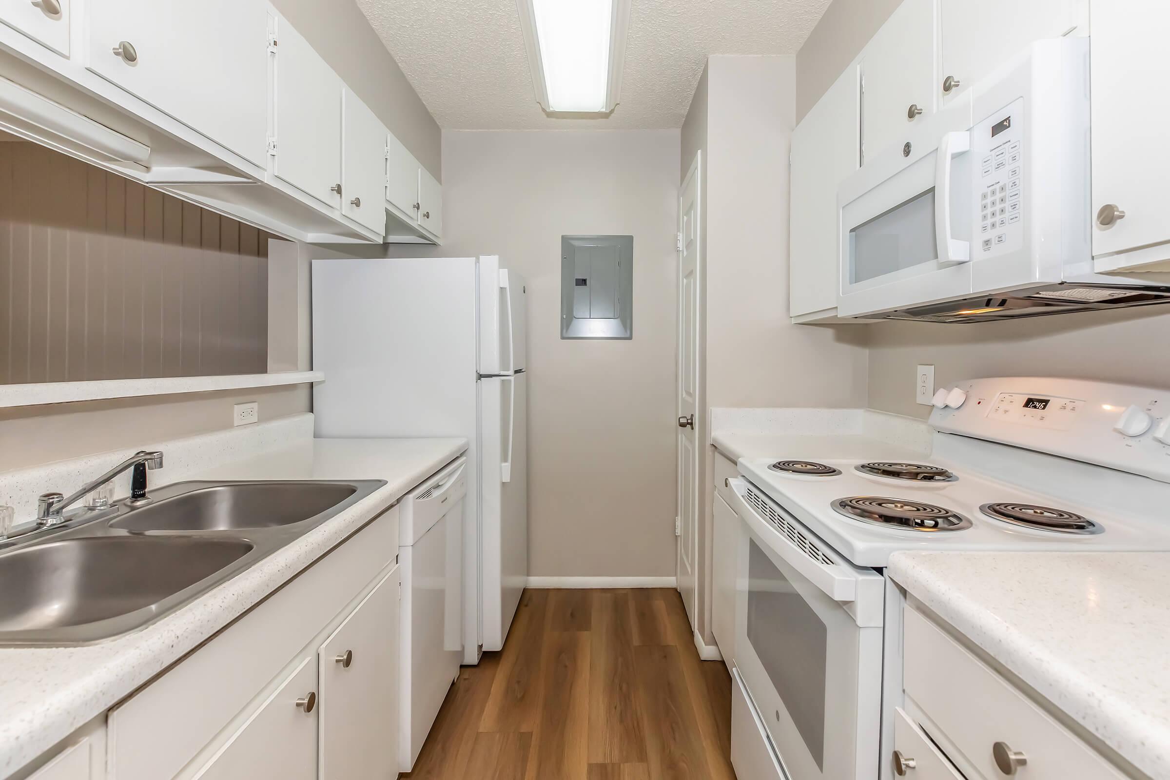 A modern kitchen featuring white cabinetry, a double sink, a white refrigerator, a microwave, and an electric stove. The space has a light color scheme with laminate flooring and ample cabinet storage, creating a clean and functional cooking area.