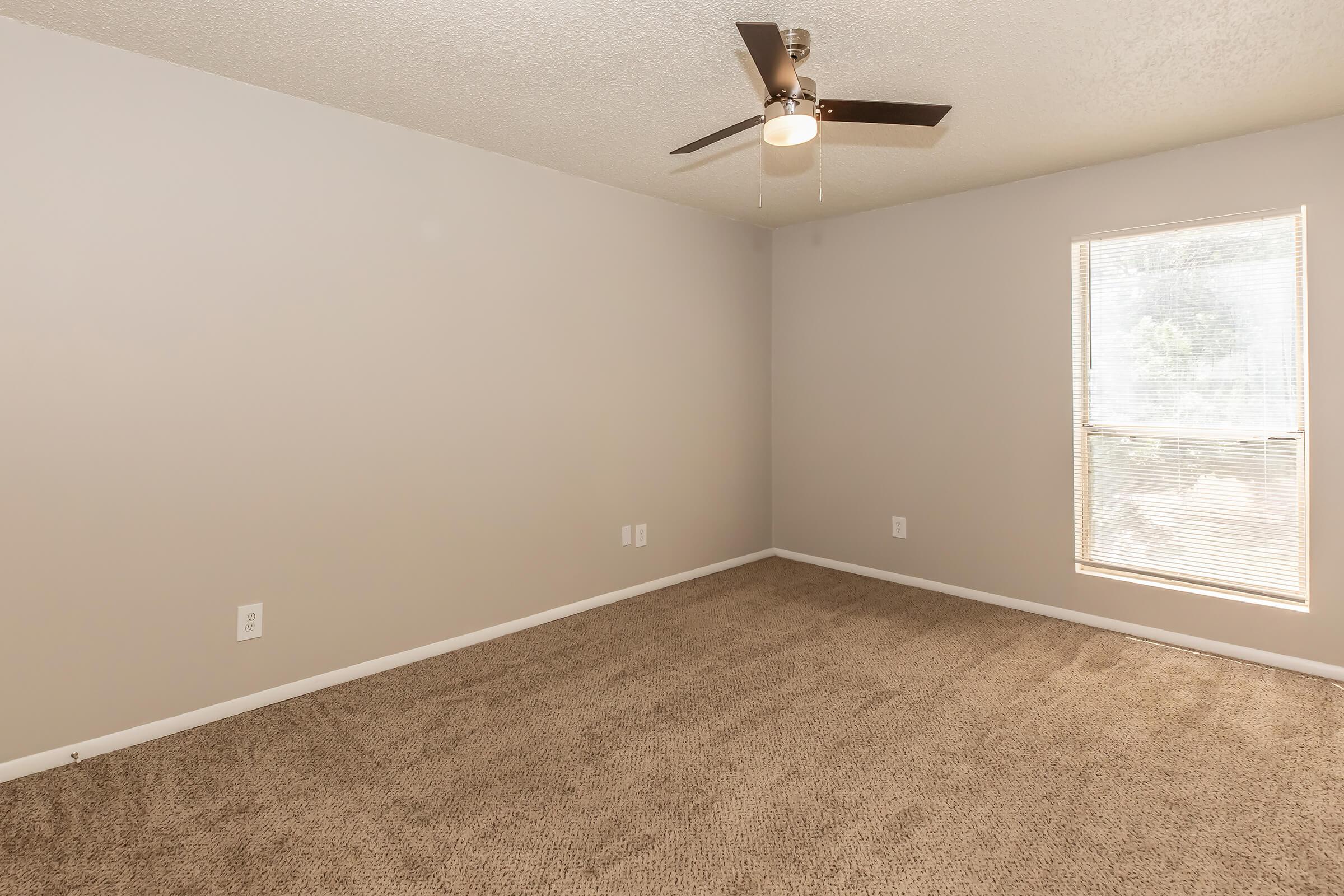 A vacant room with beige carpet and light gray walls. A ceiling fan is installed, and there is a window allowing natural light in. The space is empty, creating a clean and neutral environment suitable for various purposes.