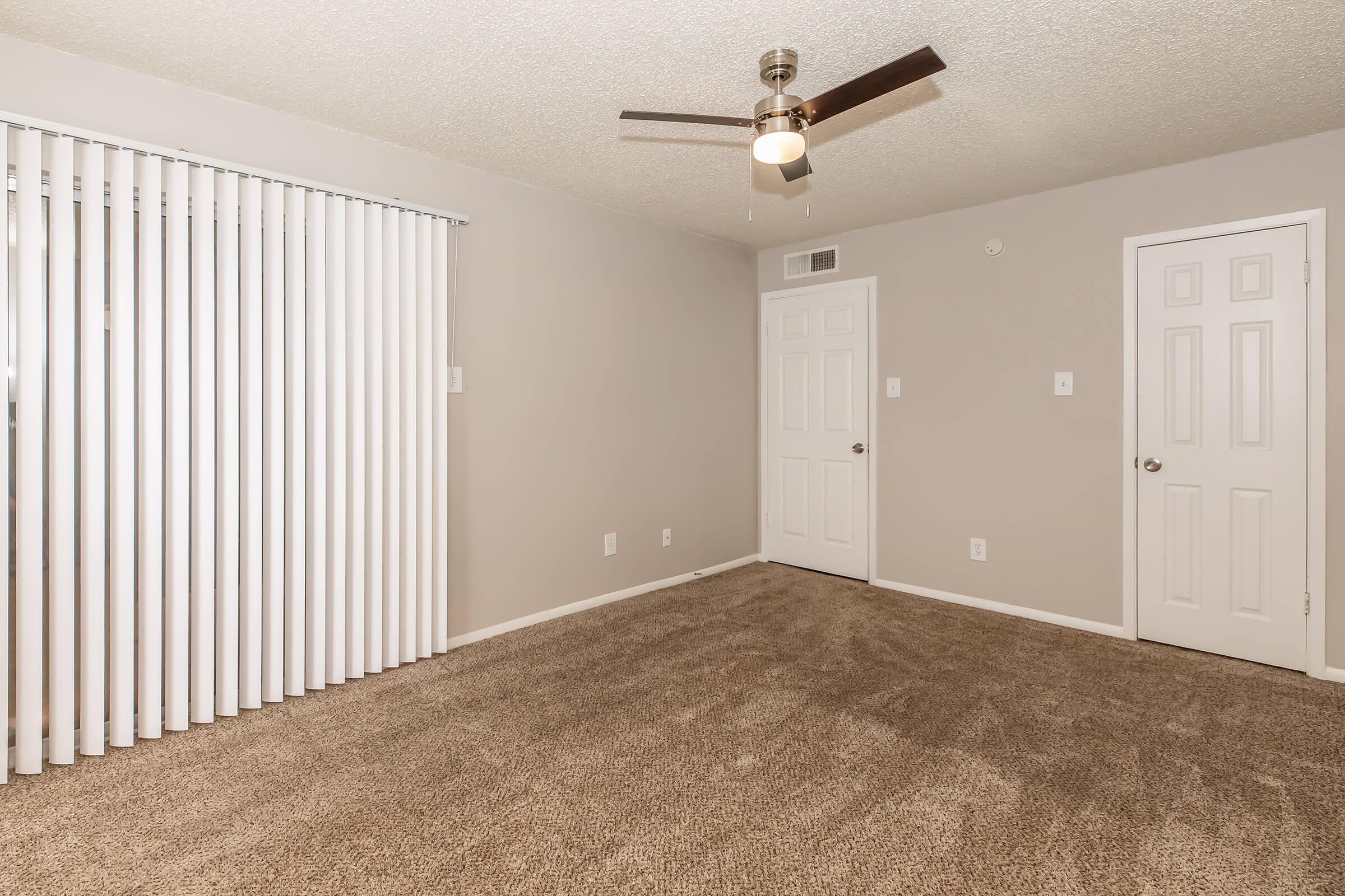 A neutral-toned empty room with beige carpet and light gray walls. It features vertical blinds on a large window, a ceiling fan, and two closed doors against the wall. The space is well-lit, creating a clean and spacious atmosphere, suitable for various furnishings and decor.