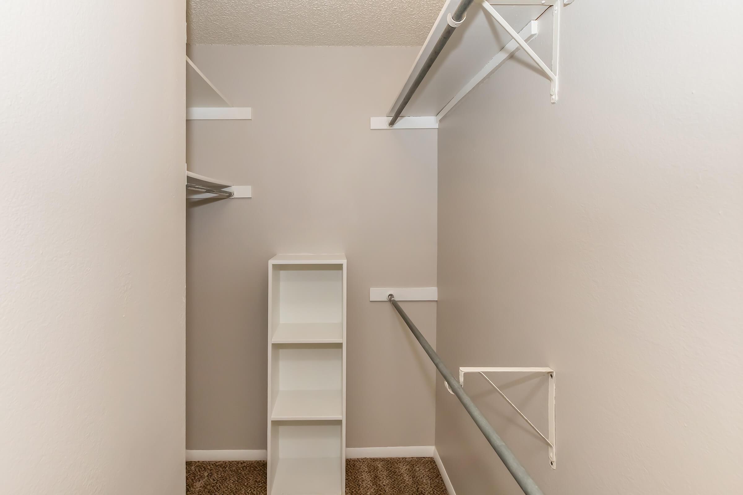 A small, empty closet featuring a light-colored wall, a carpeted floor, and two hanging rods. One side includes a white shelf unit, providing storage space. The overall design is simple and functional, ideal for organizing clothes and accessories.