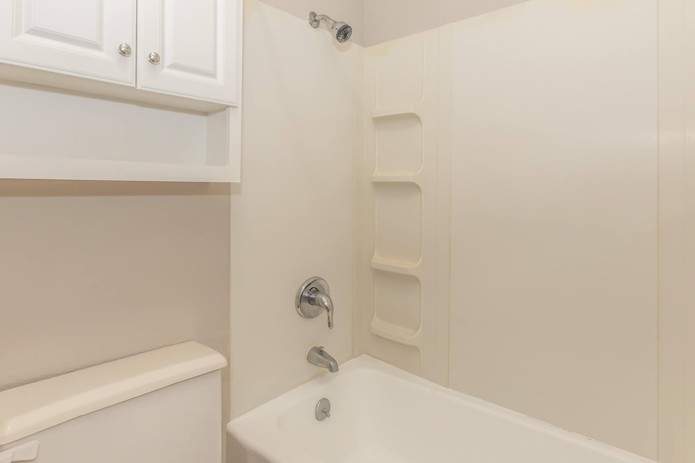 A clean and modern bathroom featuring a white bathtub with a wall-mounted showerhead, a simple faucet, and a white toilet. Above the tub is a shelf unit with white cabinetry providing additional storage. The walls are a light color, enhancing the space's brightness and cleanliness.