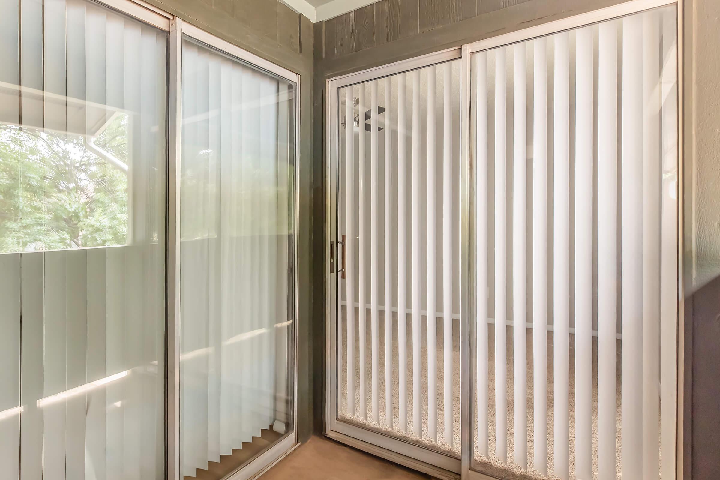 A sunlit room featuring large glass sliding doors with vertical blinds. The walls are painted in a neutral color, and there's a carpeted floor. The space appears airy and bright, with greenery visible outside the doors, creating a serene indoor setting.