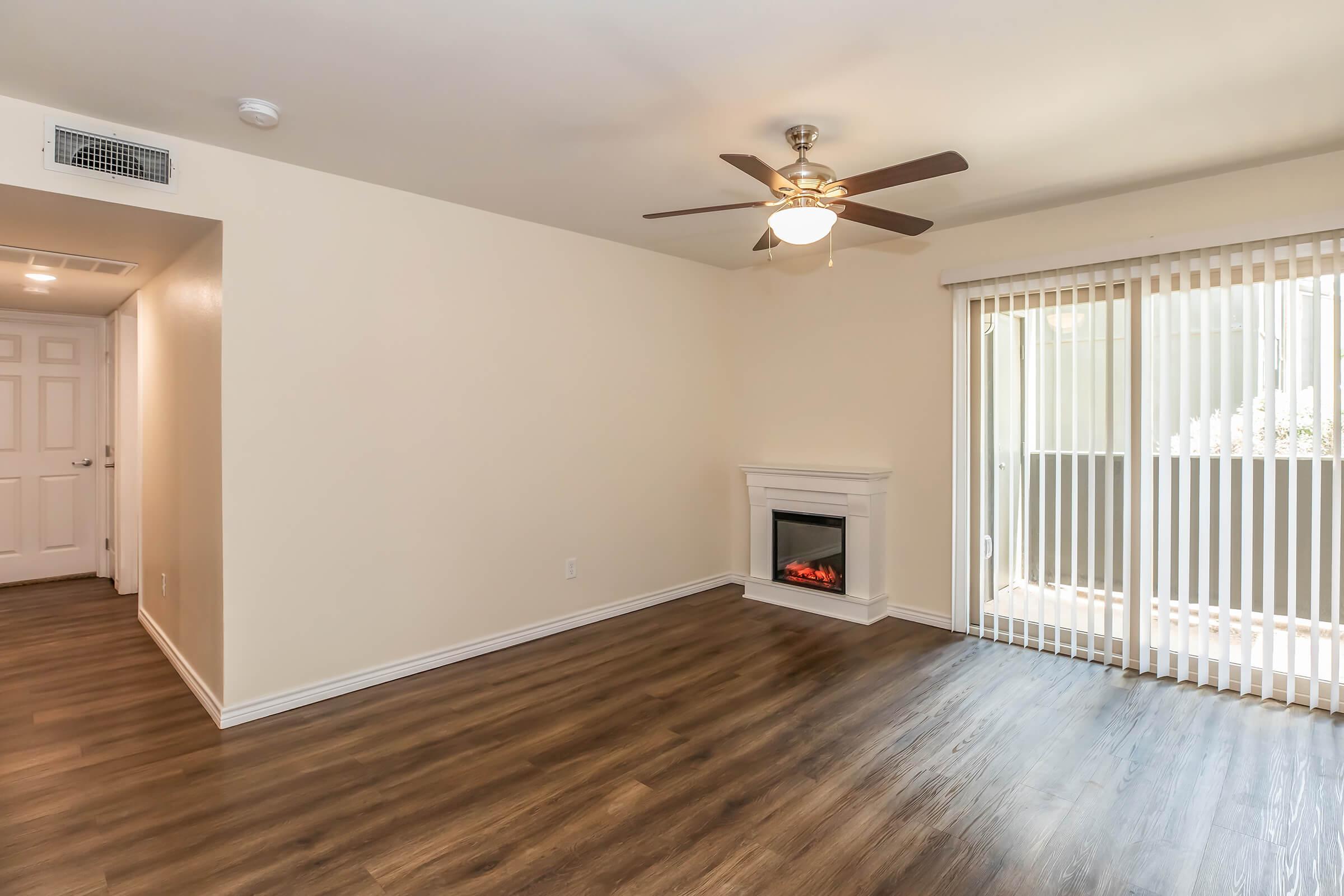 A spacious, light-colored living room featuring a ceiling fan, a faux fireplace, and large sliding glass doors with vertical blinds. The floor is made of wood-look laminate, and there’s a neutral-colored wall with a door to the left leading to another area. The overall ambiance is bright and inviting.