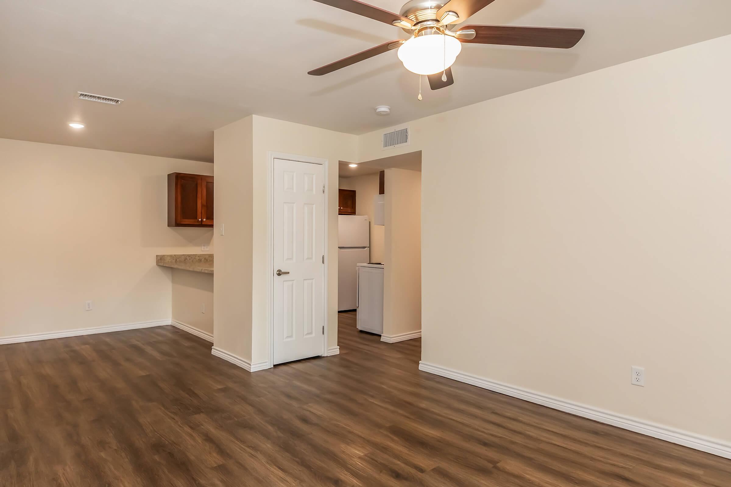 A spacious, light-filled living area featuring wood laminate flooring, a ceiling fan, and beige walls. A white door leads to a kitchen area, partially visible with light-colored cabinets. The room is uncluttered, creating a welcoming atmosphere.