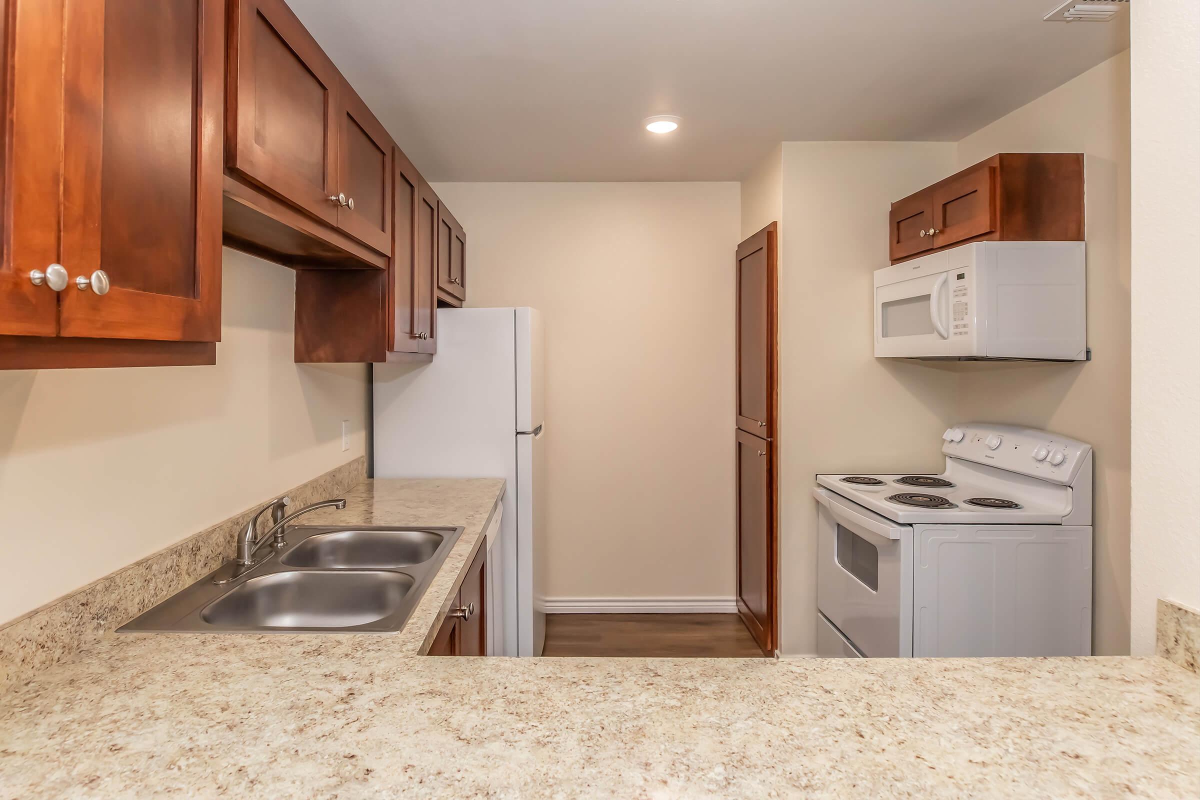A modern kitchen featuring wooden cabinets, a double sink, a white refrigerator, an oven with a stovetop, and a microwave. The walls are painted in a light color, and the countertop is made of a light-colored stone. The space is well-lit with overhead lighting.