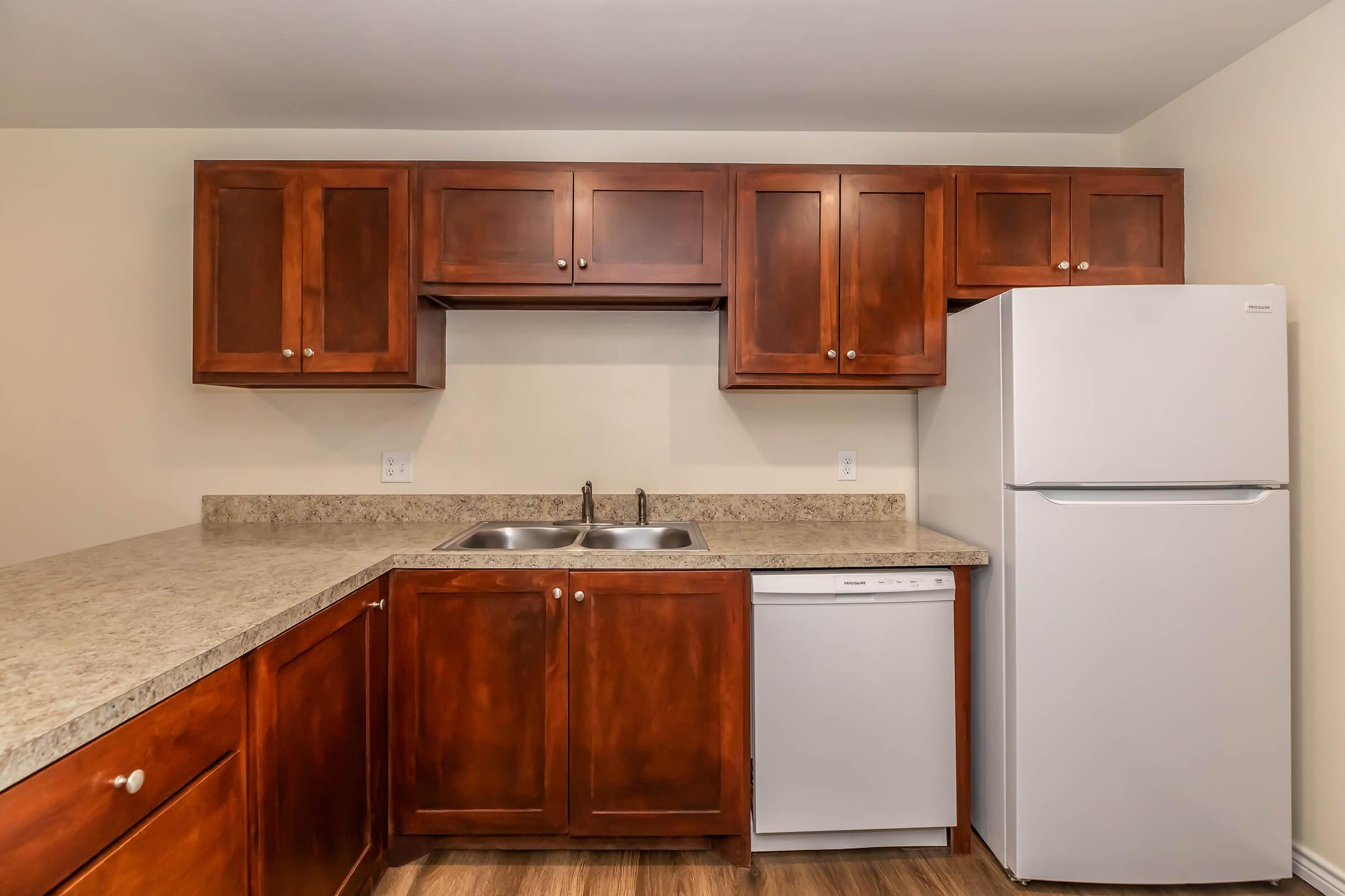 A modern kitchen featuring wooden cabinets, a granite countertop, a double sink, a dishwasher, and a white refrigerator. The walls are painted light, creating a bright and inviting atmosphere. The flooring is wooden laminate, providing a warm contrast to the cabinetry.
