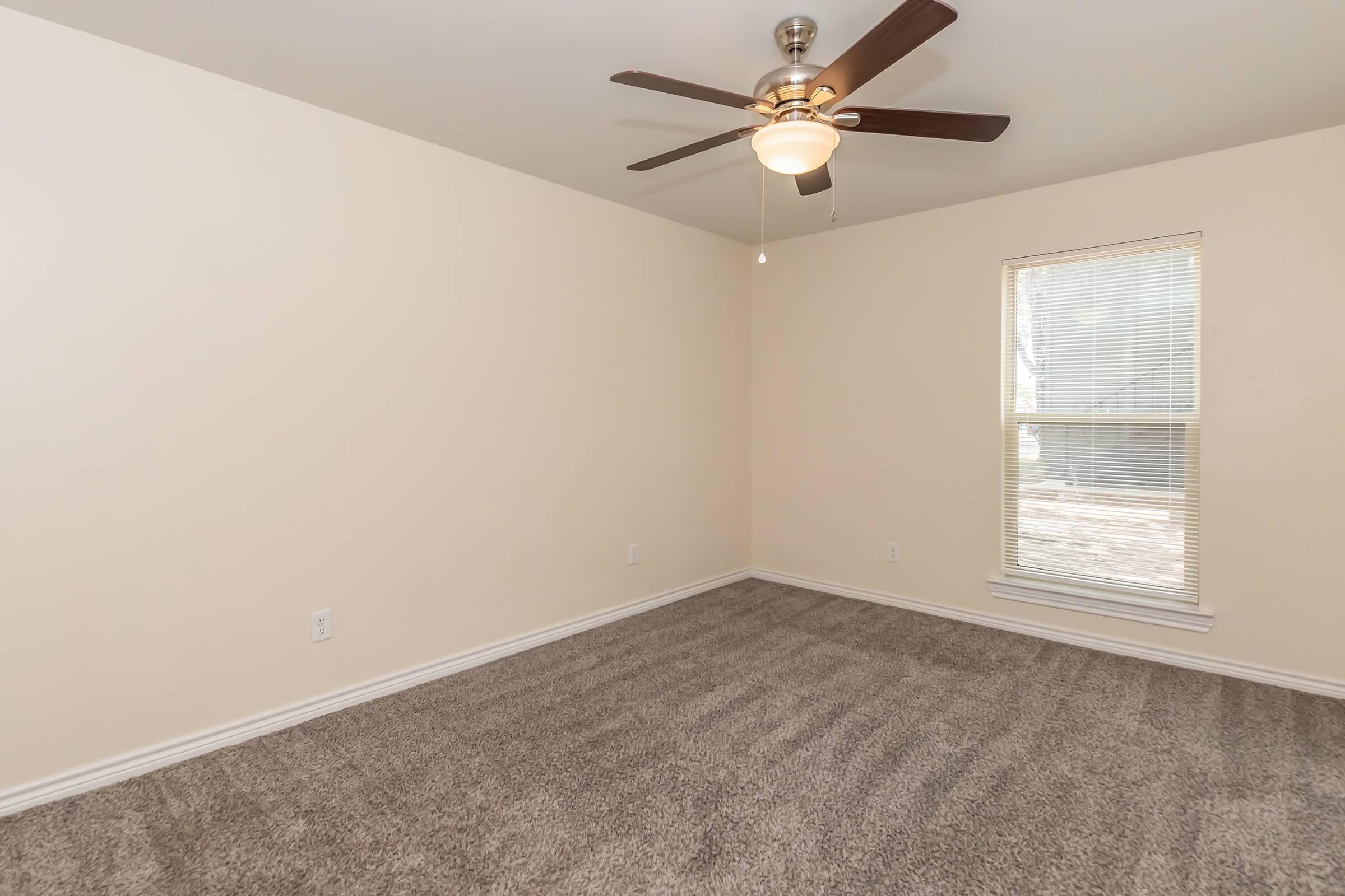 Empty room with light beige walls and a ceiling fan. The floor is covered in textured gray carpet. A window on the right lets in natural light, showcasing blinds. The room appears spacious and clean, suitable for various uses like a bedroom or office.