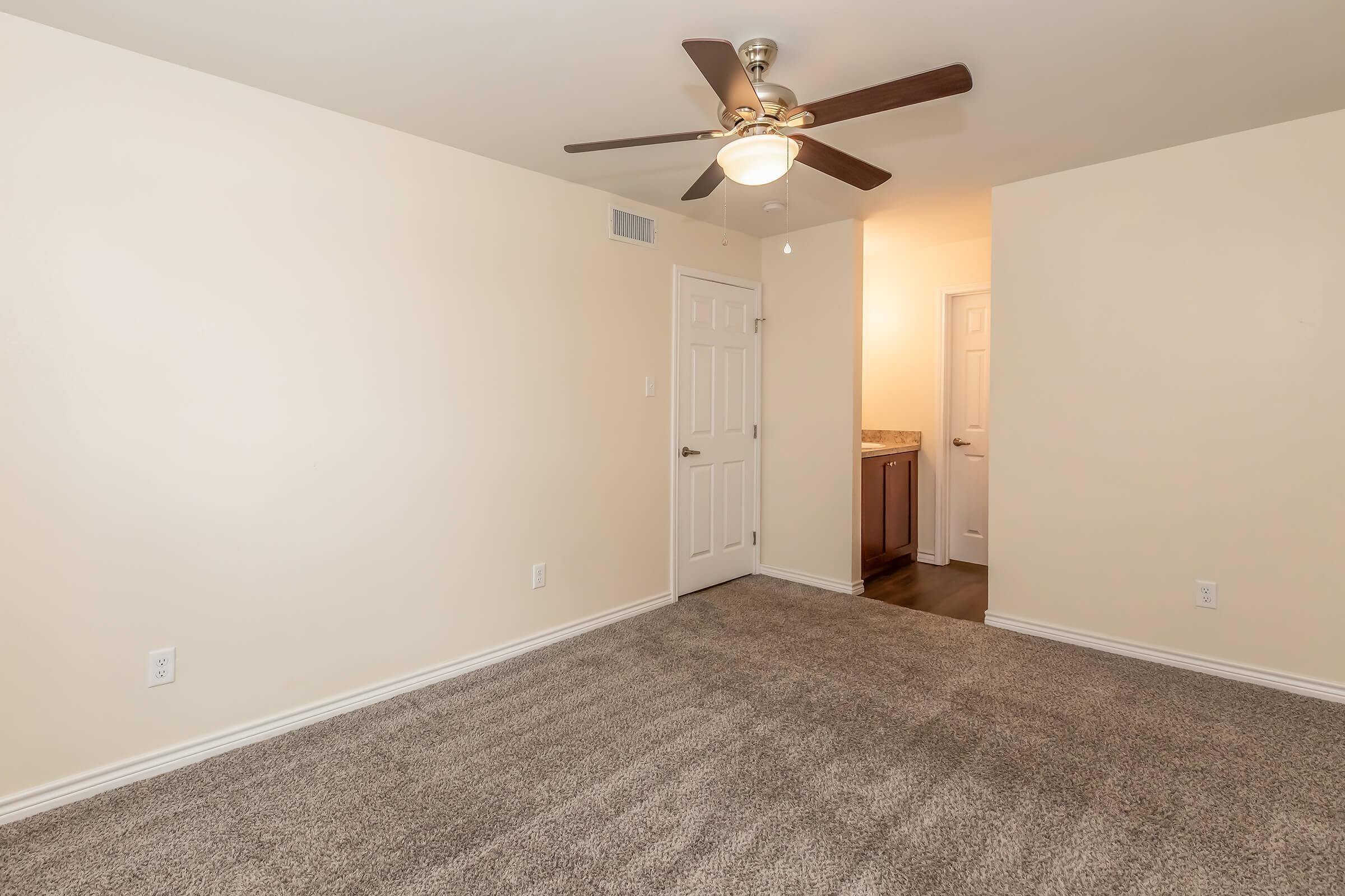 A spacious, empty room featuring beige walls and a light carpeted floor. A ceiling fan with wood blades is mounted above. To one side, a doorway leads to a small bathroom area with a dark wooden vanity visible. Natural light enters from the hallway at the back of the room.