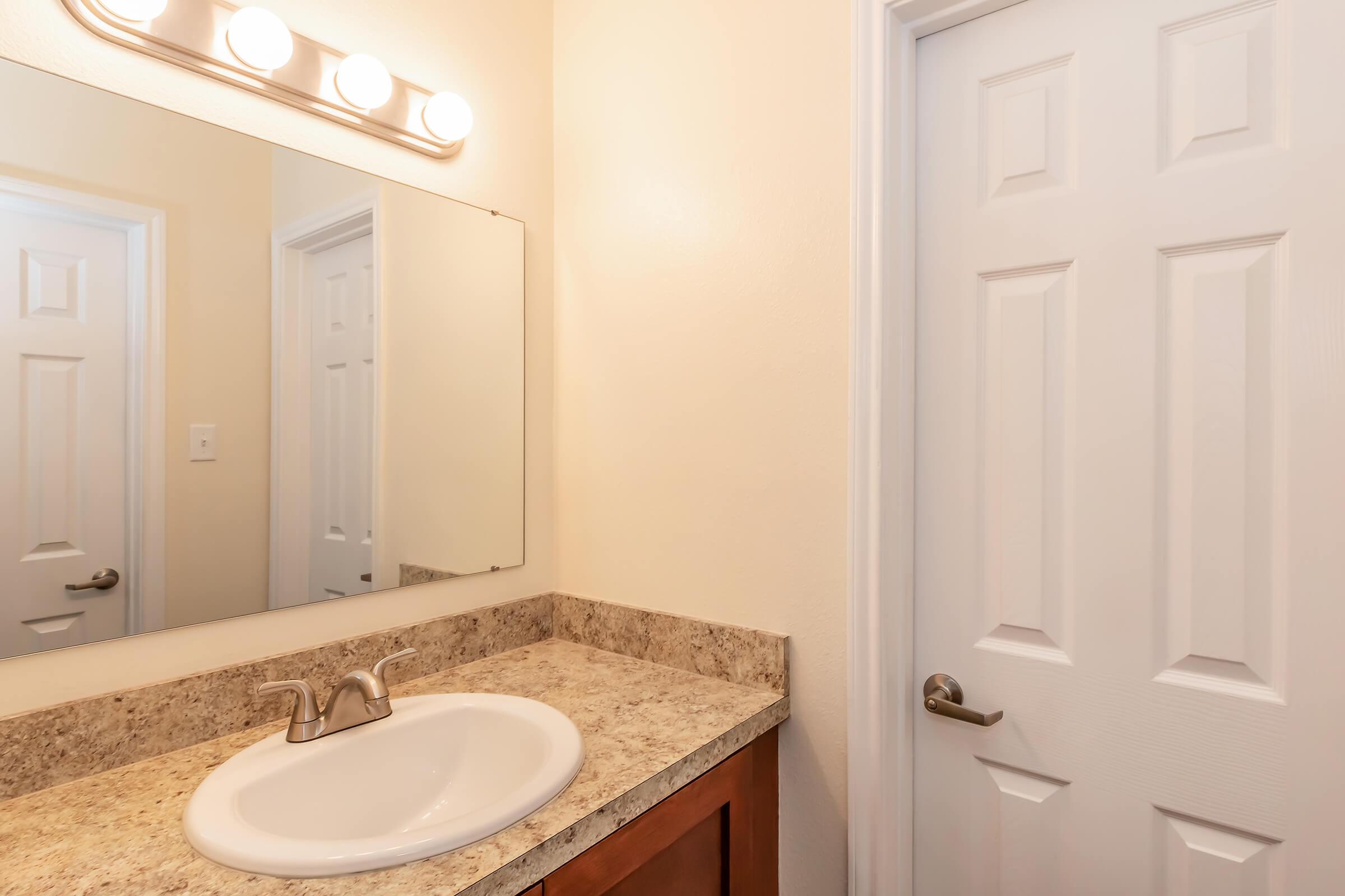 A bathroom interior featuring a sink with a curved basin, a mirror above it, and a light fixture with four bulbs. The wall is painted a light color, and there is a door on the right side, leading to another room. The countertop is made of a speckled beige stone.