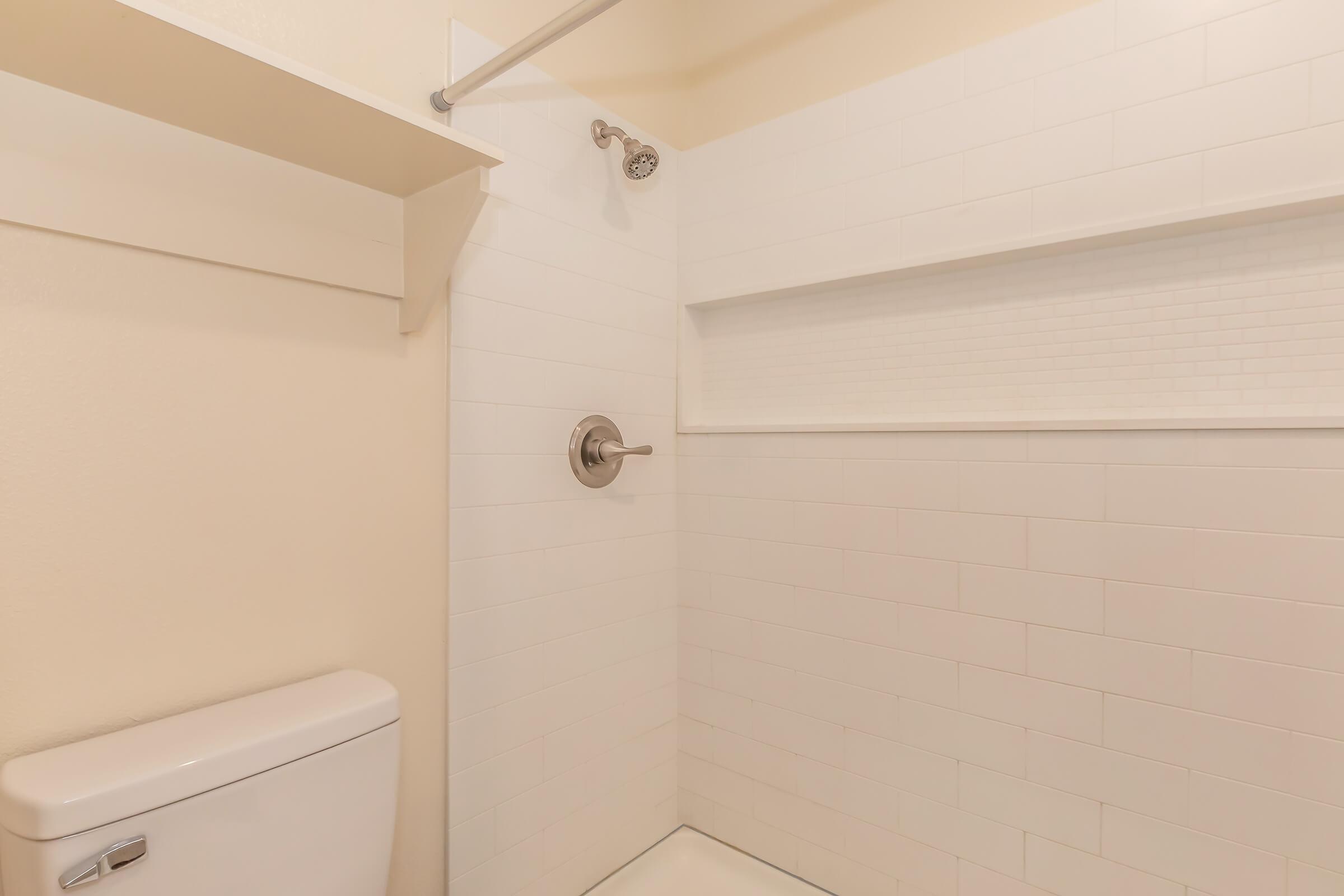 A clean, modern bathroom with a white tiled shower area. The shower features a stainless steel showerhead and a built-in shelf. A white toilet is visible nearby, against light beige walls, creating a bright and spacious appearance.