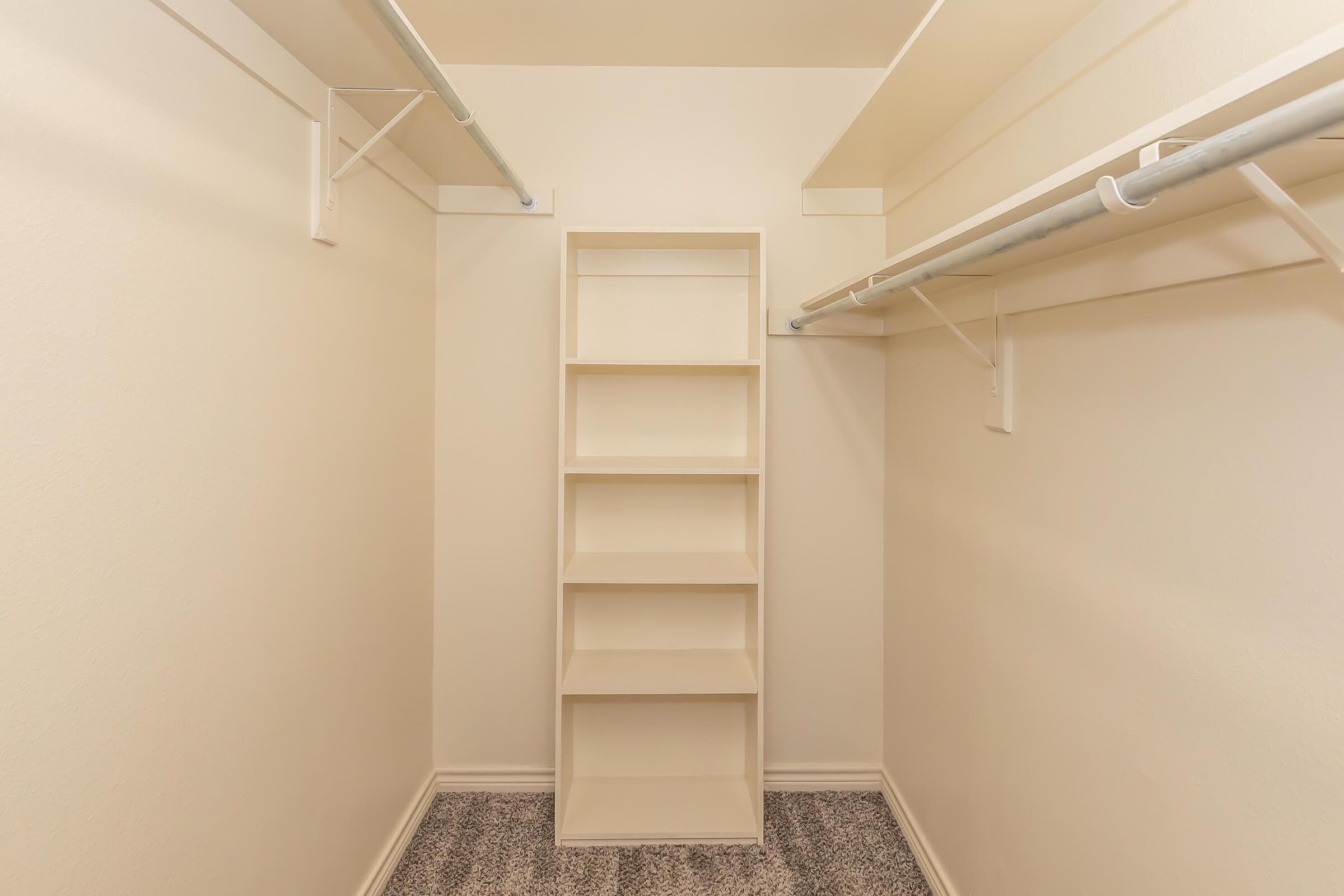 A clean, empty walk-in closet featuring light-colored walls and carpet. It includes two hanging rods on either side for garments and a central shelving unit with five shelves for storage. The overall space appears organized and spacious, ready for storing clothes and other items.