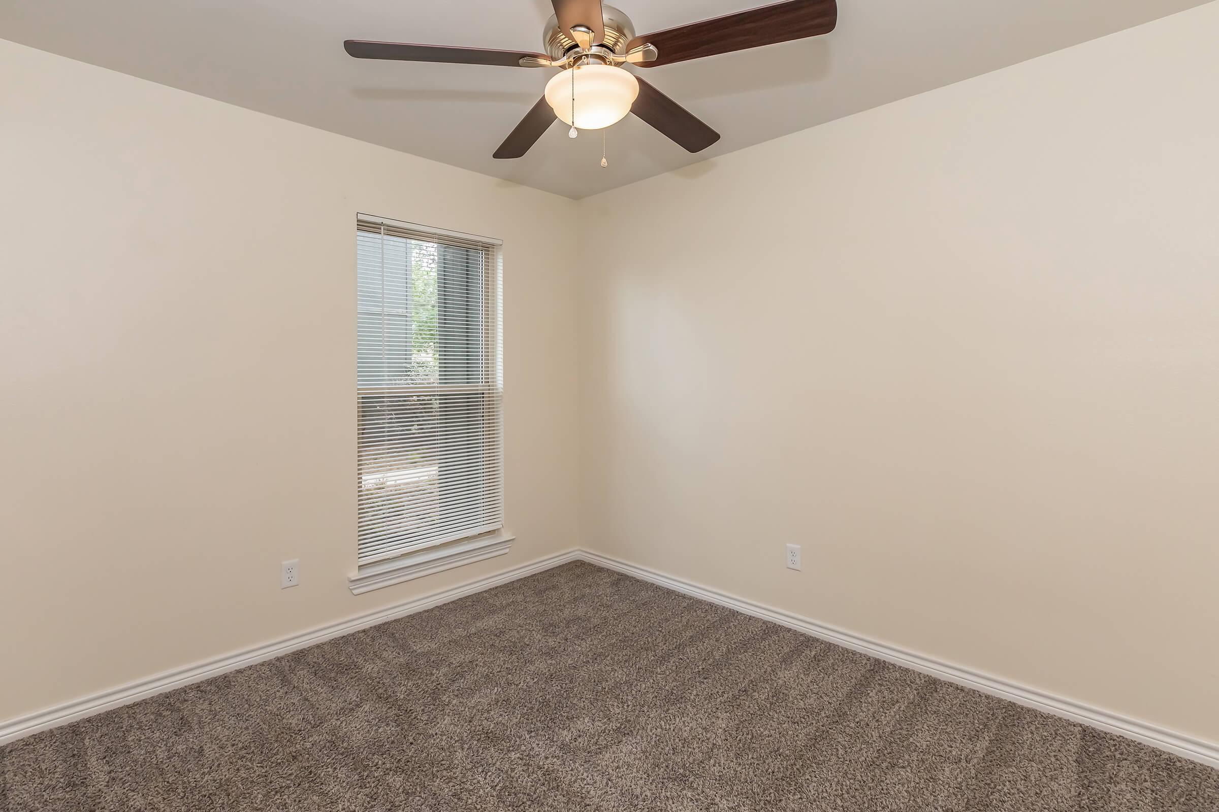 Interior of a vacant room featuring light beige walls, a ceiling fan with light, and a window with blinds. The floor is covered in carpet, and the room is well-lit with natural light. The space appears clean and ready for new furnishings.