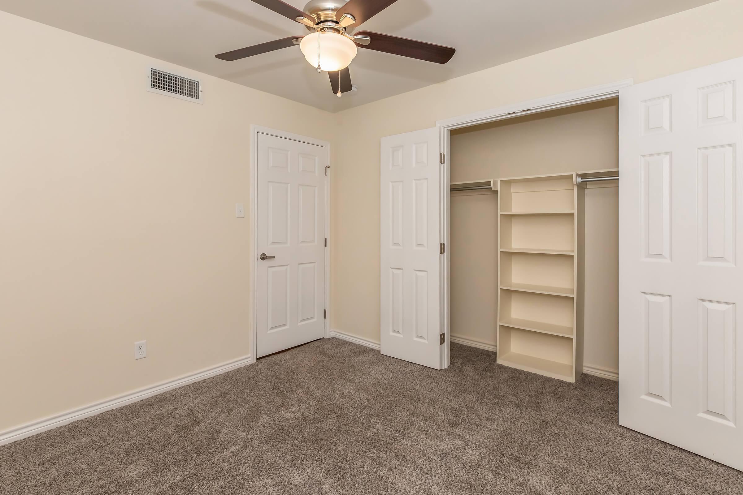 A room with light beige walls and carpet, featuring a ceiling fan with a light fixture. Two white doors are visible: one leads to a closet with shelves, and the other is closed. The room is spacious and well-lit, creating a neutral aesthetic.