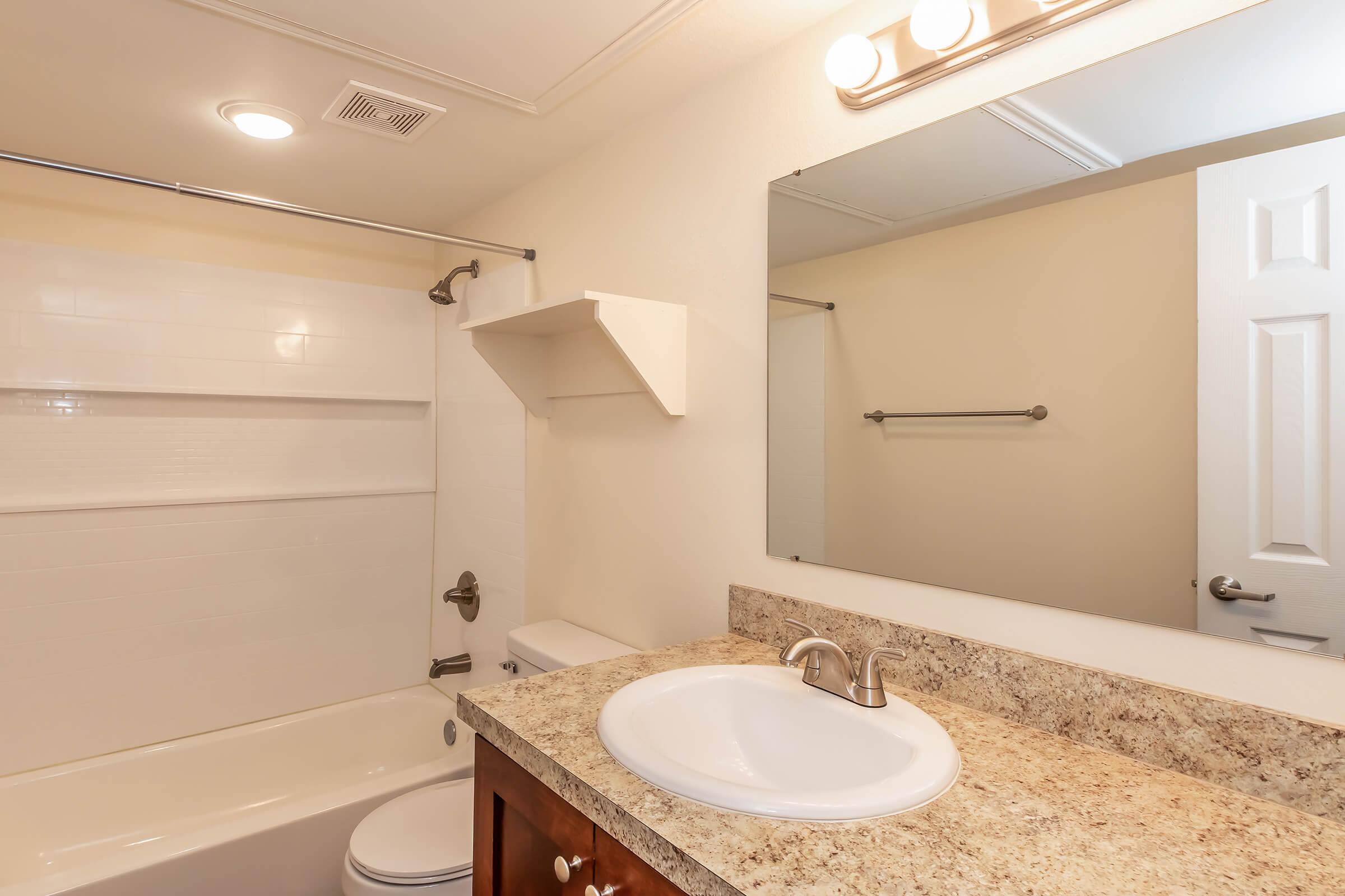 A clean and modern bathroom featuring a white bathtub with a shower, light-colored walls, a sleek countertop with a sink, and a large mirror. There is a towel bar and a small shelf for storage, creating a neat and functional space.