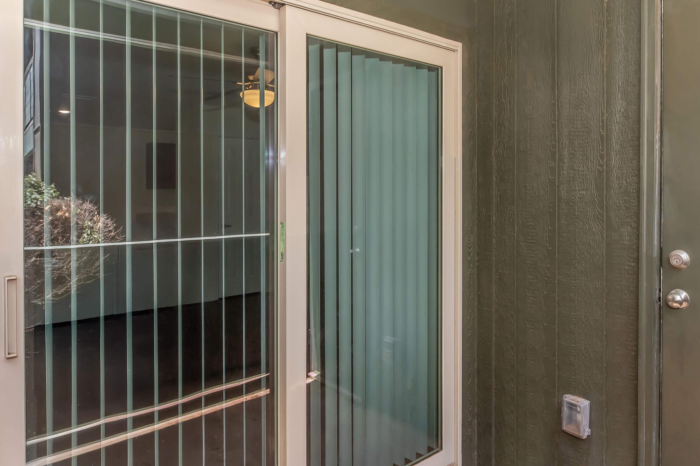 A glass door with vertical blinds, leading to a room inside a building. The exterior walls are painted a dark green, and a small panel next to the door contains a doorknob and a security feature. The interior is partially visible through the door, suggesting an open space beyond.