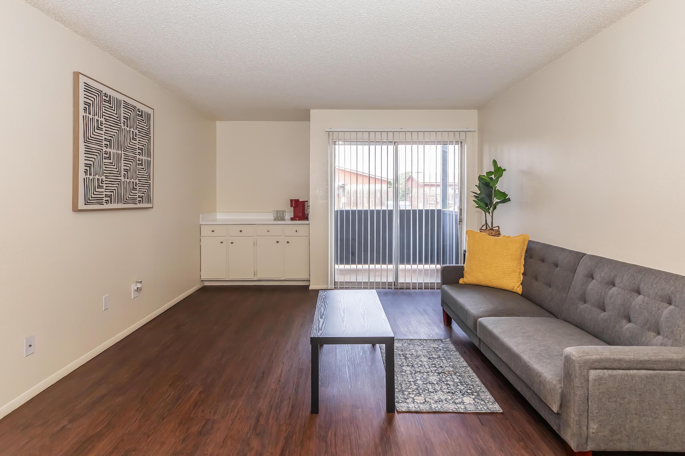 A modern living room featuring a gray sofa with a yellow throw pillow, a wooden coffee table, and a patterned rug. There's a minimalist wall art piece on the left, and large sliding glass doors leading to a balcony, allowing natural light to fill the space.