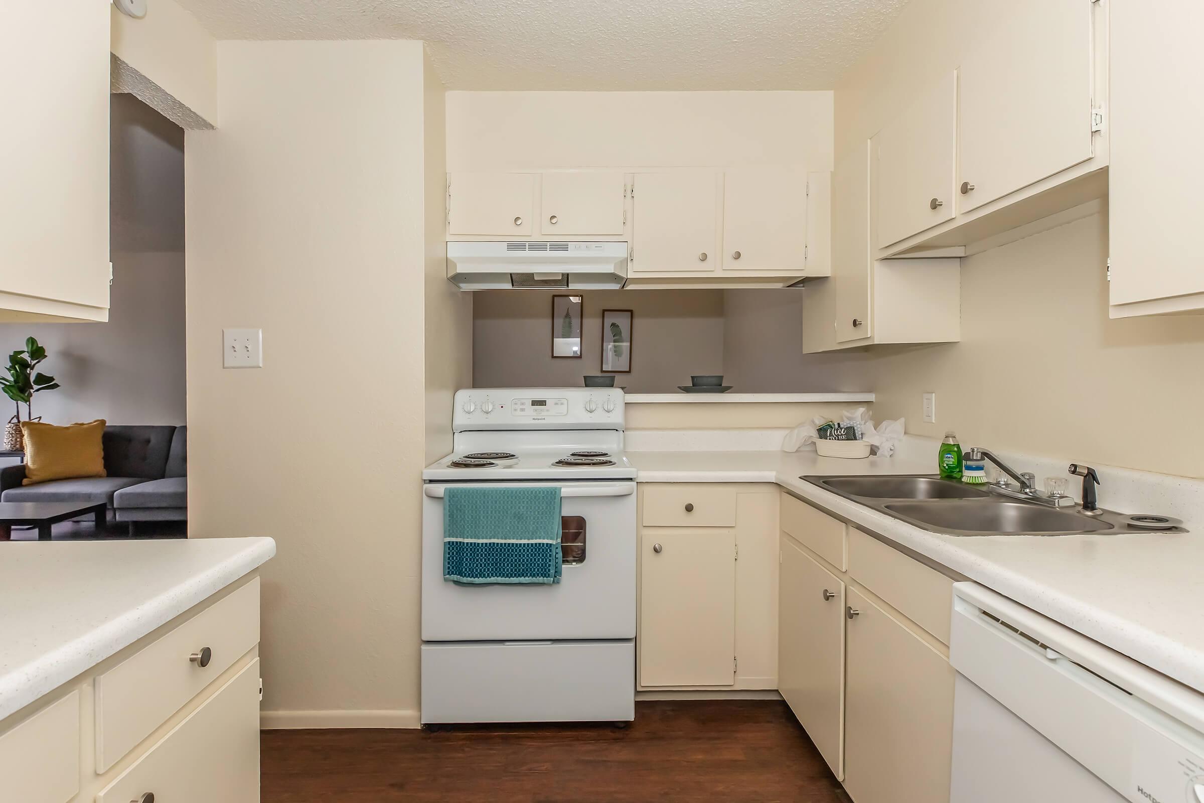 A clean and modern kitchen featuring white cabinets, a white stove with an overhead range hood, a sink, and a countertop. A teal towel hangs on the stove. The kitchen has a neutral-colored wall and a glimpse of a living area with a couch in the background.