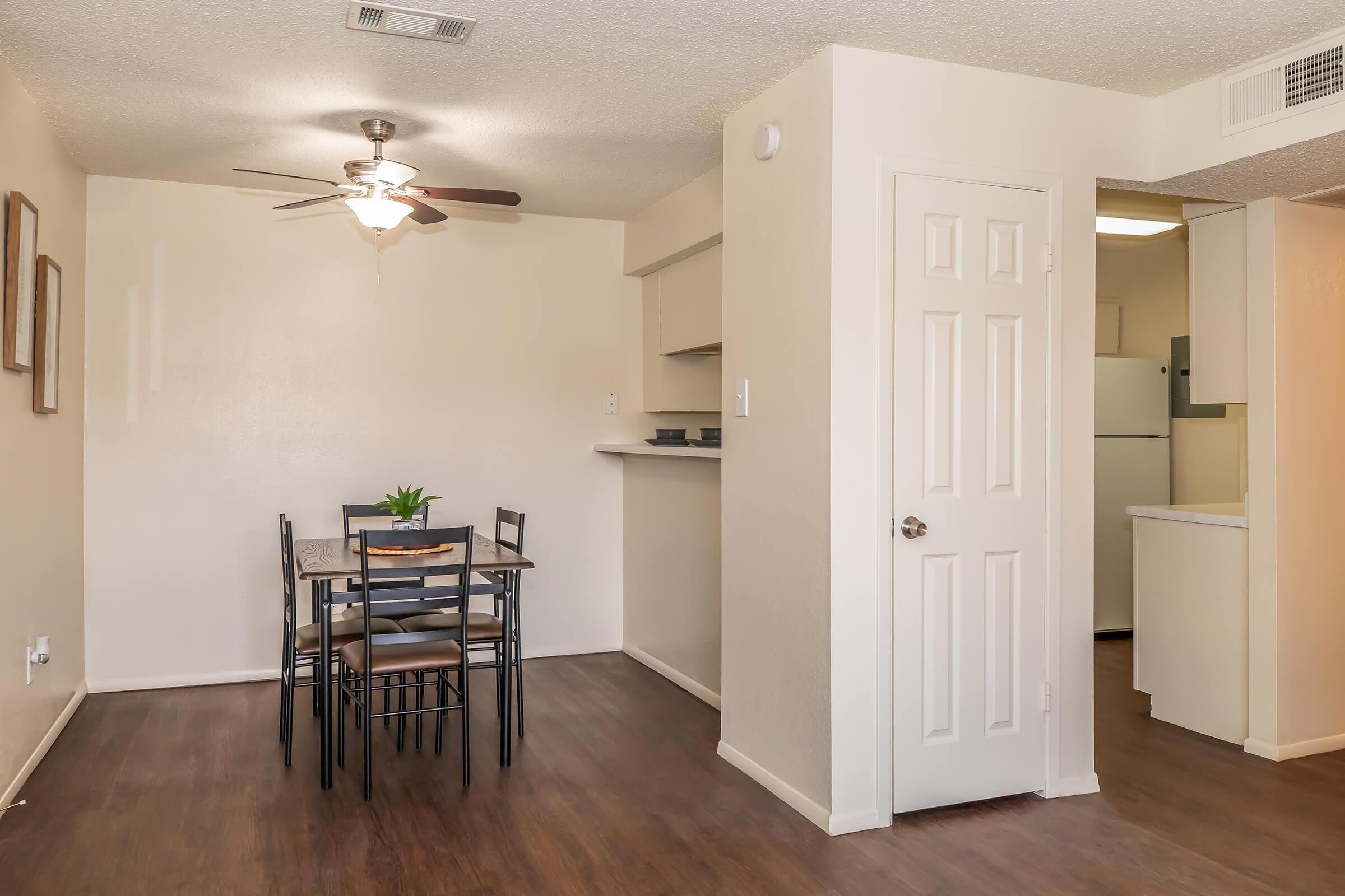 A small dining area with a table and chairs, a ceiling fan above, and light-colored walls. A doorway leads to a kitchen area, partially visible, and there's a potted plant on the table. The floor is a dark wood finish, creating a cozy atmosphere.