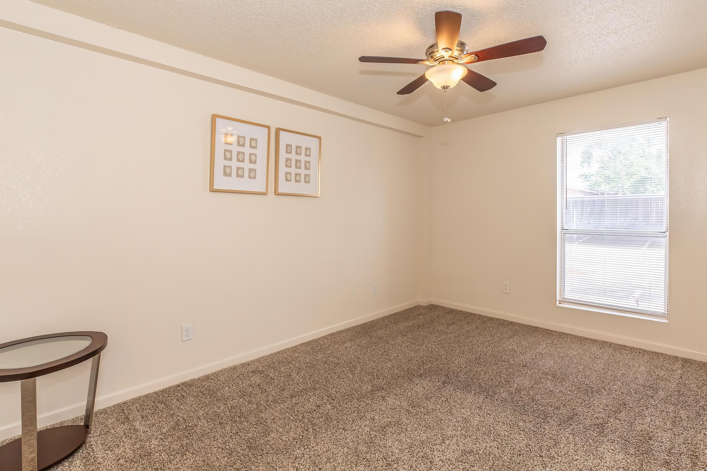 A spacious, well-lit room with beige walls and a ceiling fan. Features a small, modern side table and two framed pictures on the wall. A large window allows natural light to brighten the space, which is carpeted in a neutral tone. Ideal for a cozy living area or bedroom.