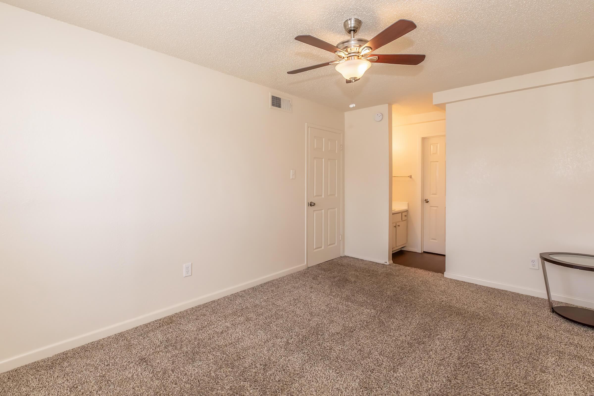 A spacious, well-lit room with beige walls and carpeted flooring. It features a ceiling fan, a door leading to a bathroom area, and an empty corner suitable for furniture placement. The overall atmosphere is clean and inviting, ideal for a living or bedroom space.