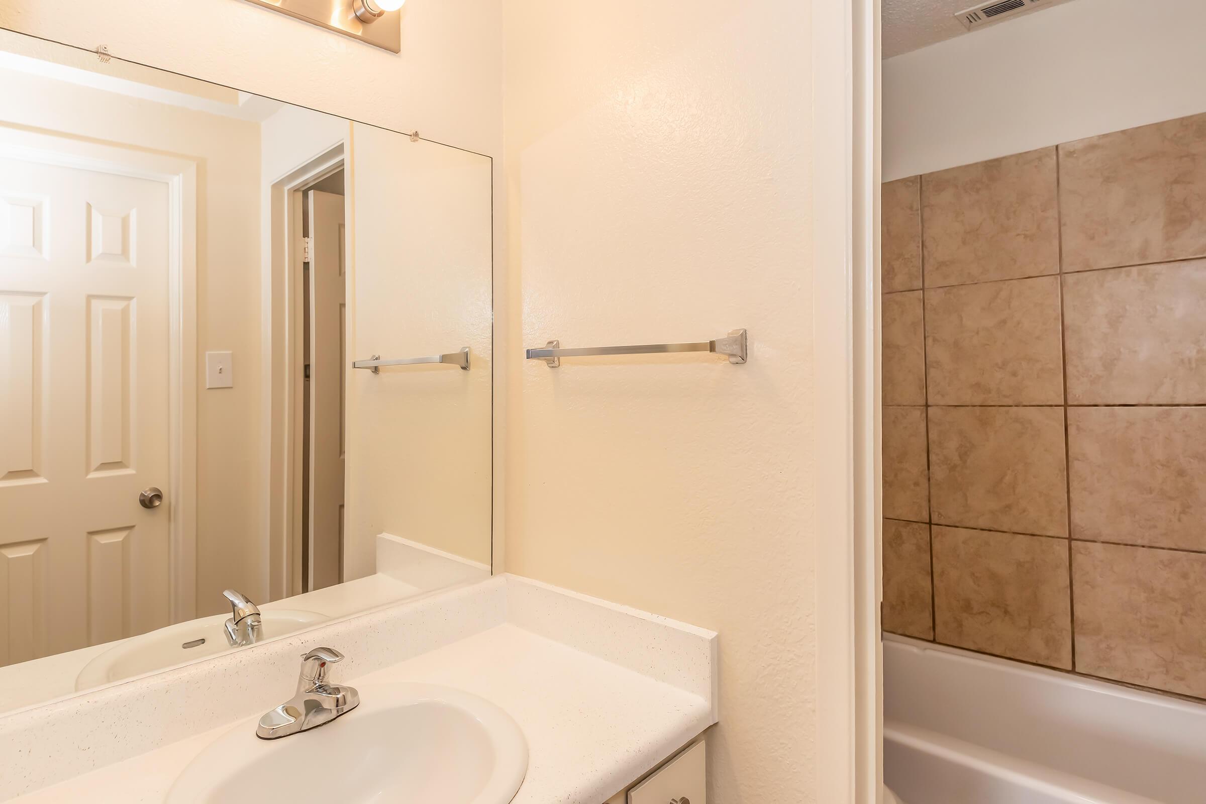 A clean bathroom with a white sink and countertop, a large mirror above the sink, and light beige walls. A shower/tub combination with tiled walls is visible to the right. There is also a towel bar mounted on the wall and a door leading to another room in the background.