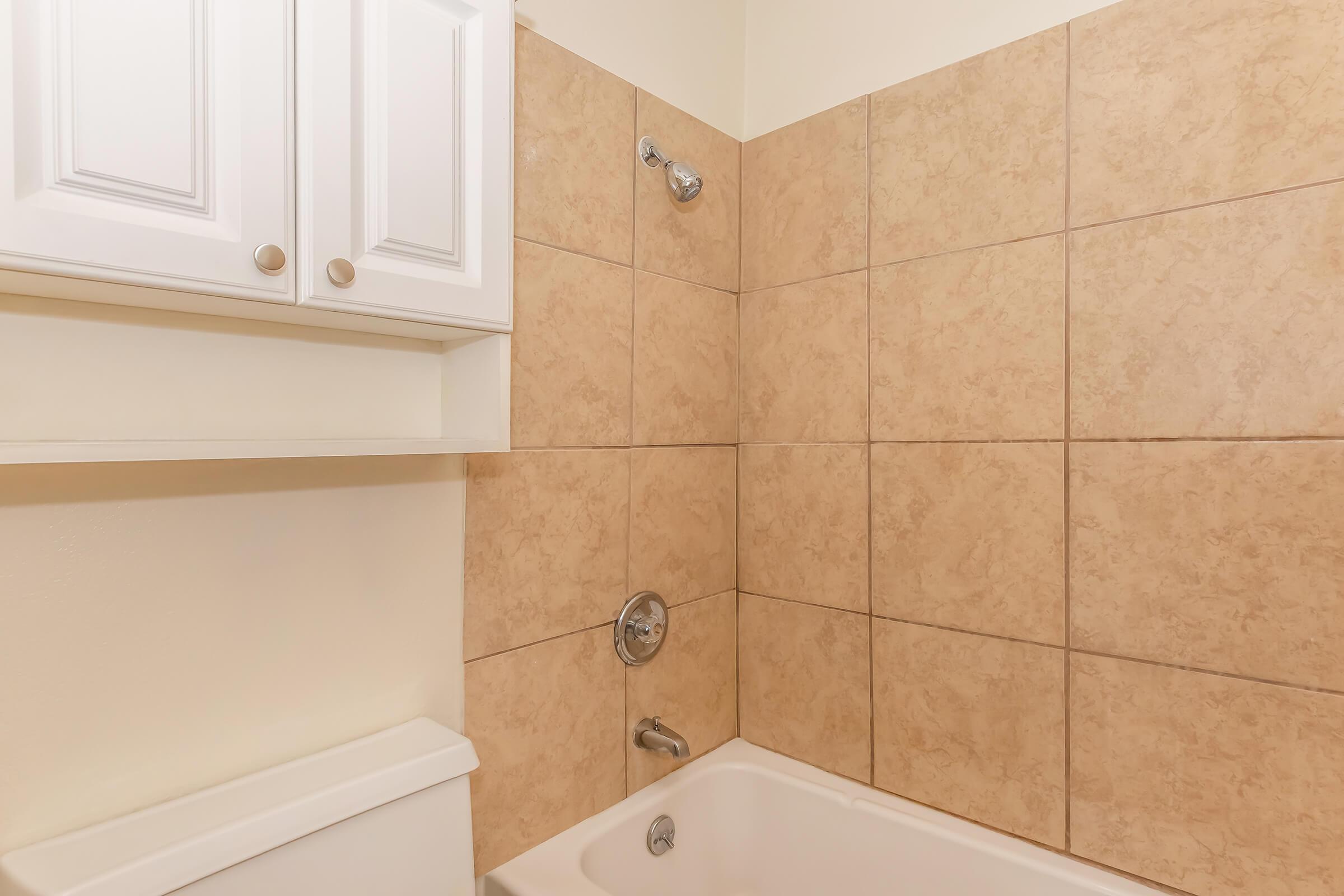 A bathroom with a beige tiled shower and bathtub. The shower features a silver faucet and a showerhead mounted on the wall. Above the tub, there's a simple white cabinet for storage, and the walls are light-colored, creating a clean, modern look.