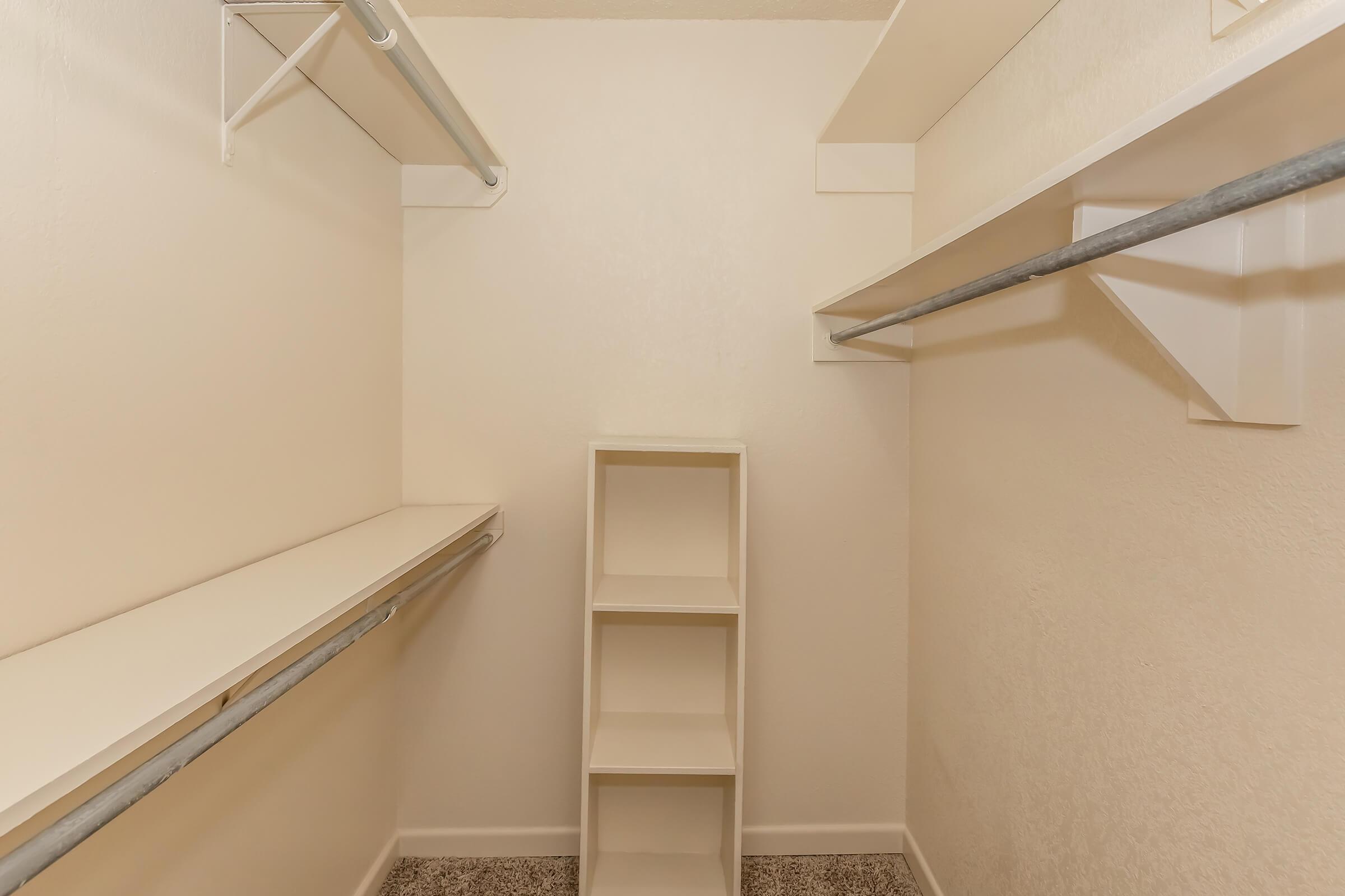 A spacious, empty closet featuring light-colored walls, two horizontal rods for hanging clothes, and a small shelving unit in the center. The floor is carpeted, and there is ample space for storage.