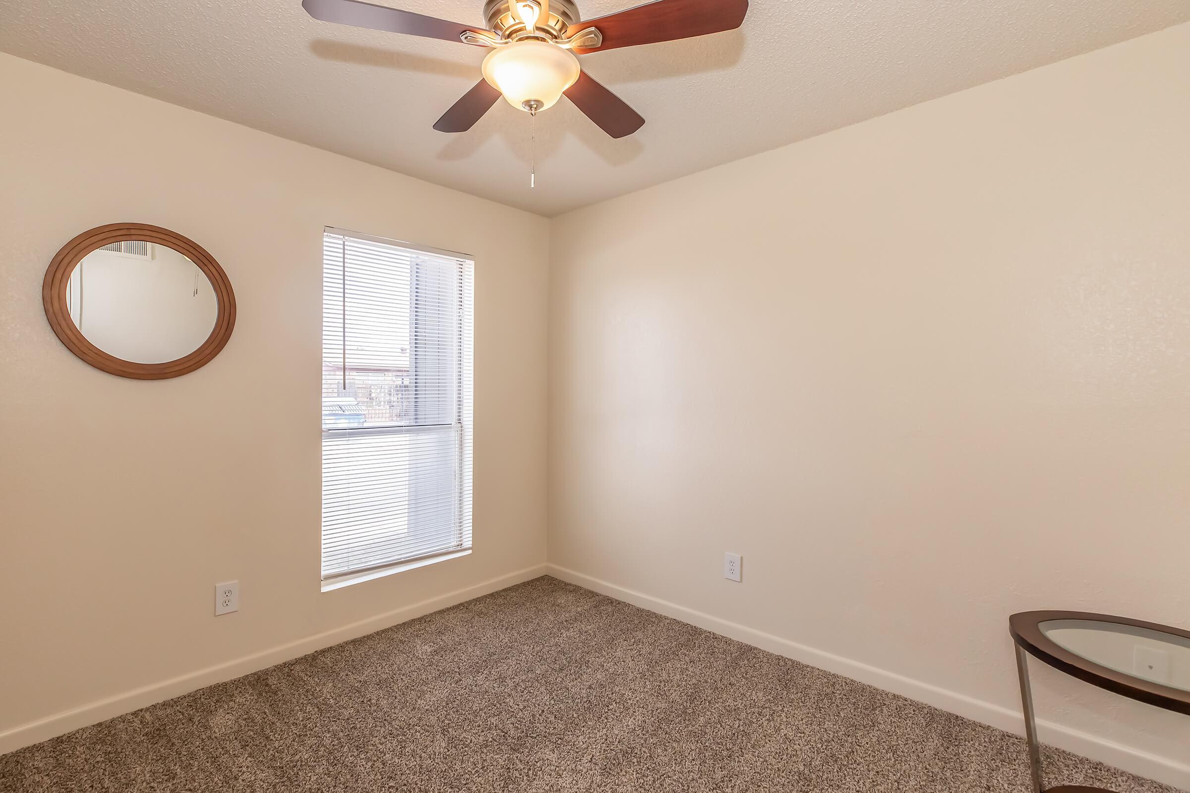 A simple, light-colored room featuring beige walls, a ceiling fan with light, and carpeted flooring. There's a round mirror on the wall, a window allowing natural light, and a small glass table against the wall. The space appears clean and uncluttered, suitable for various uses.