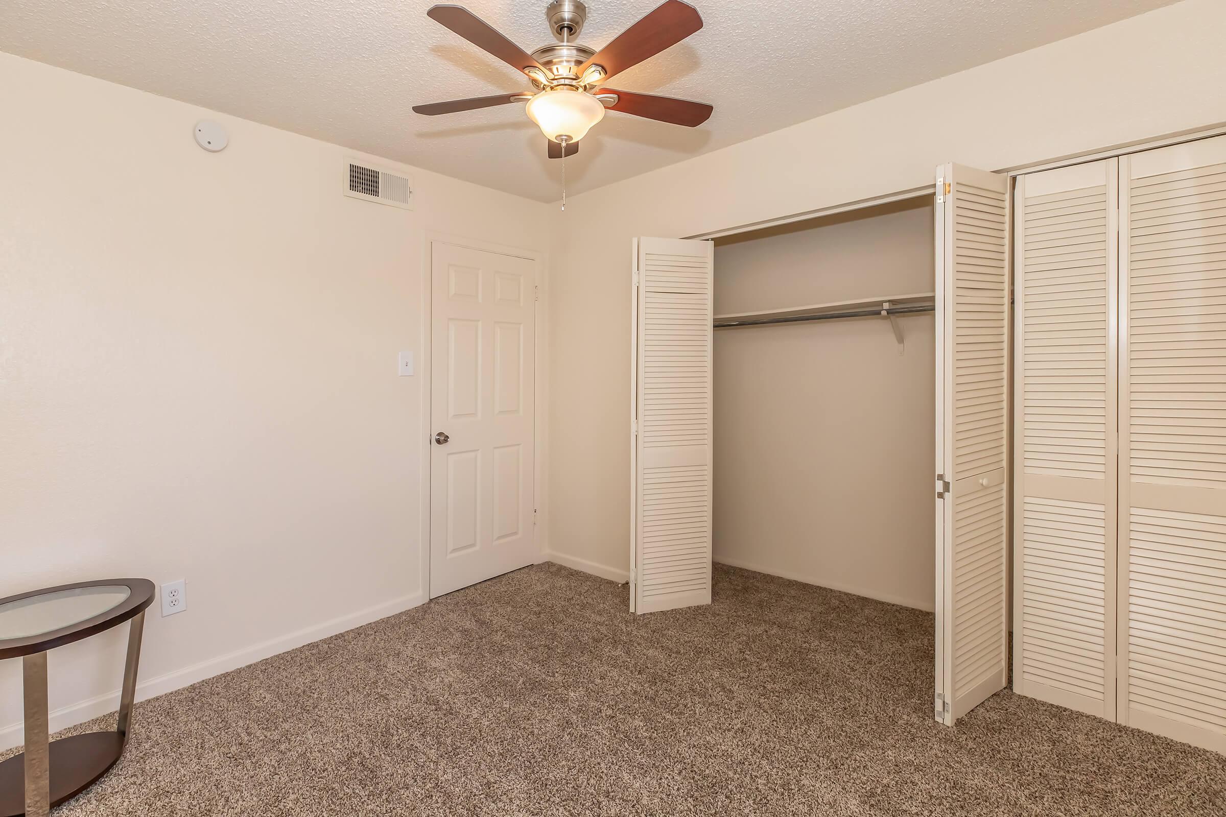 Interior view of a room featuring a ceiling fan with wooden blades, a light fixture, and beige walls. There is a small round table to the left and a closet with bifold doors on the right, which is empty and has a single hanging rod. The floor is covered with carpet in a neutral tone.
