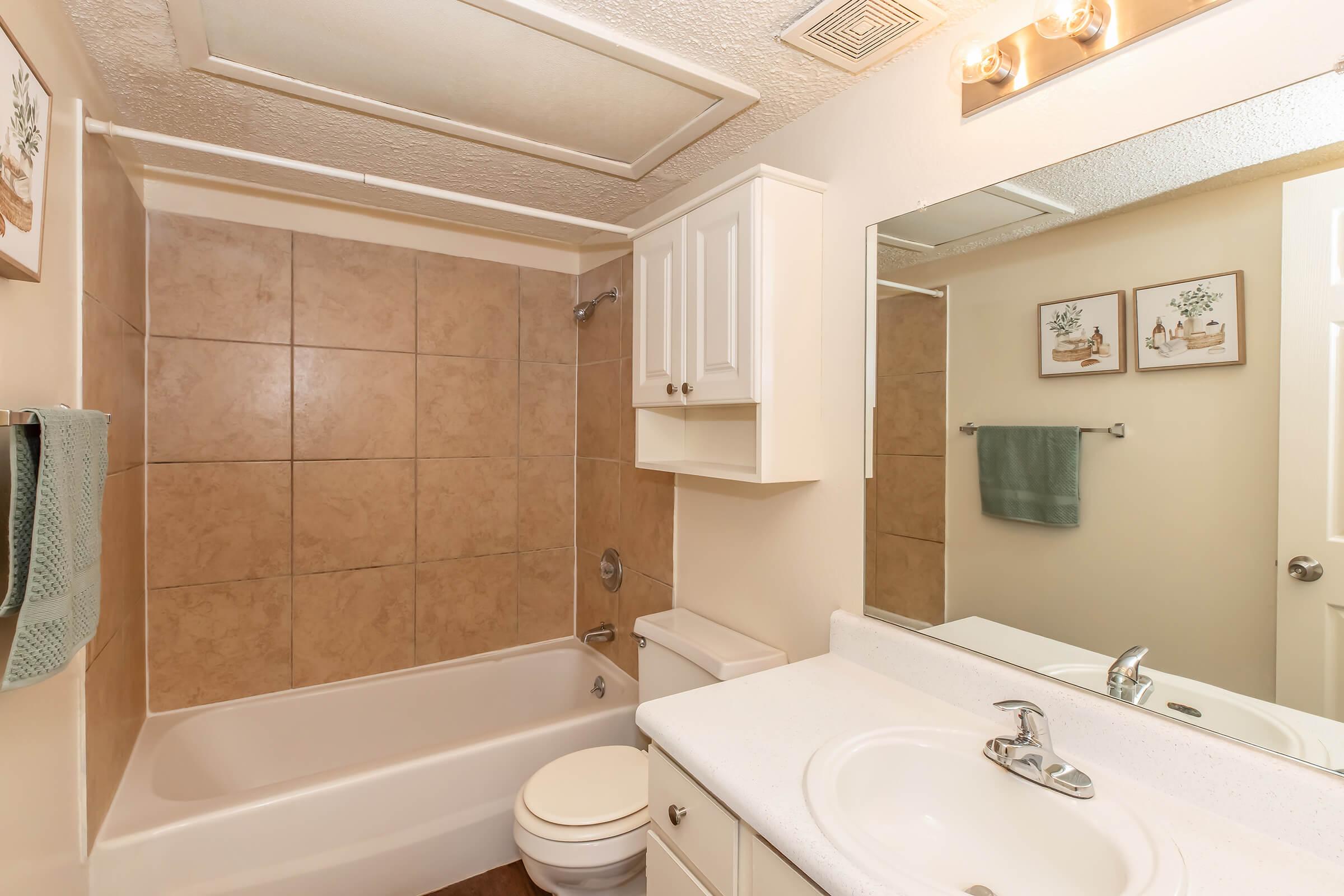 Modern bathroom featuring a beige tiled shower/bathtub, white vanity with sink, and wall-mounted mirror. Shelves above the toilet and decorative wall art complete the design. Soft lighting from four fixtures above the mirror creates a warm ambiance. Space is clean and well-organized.