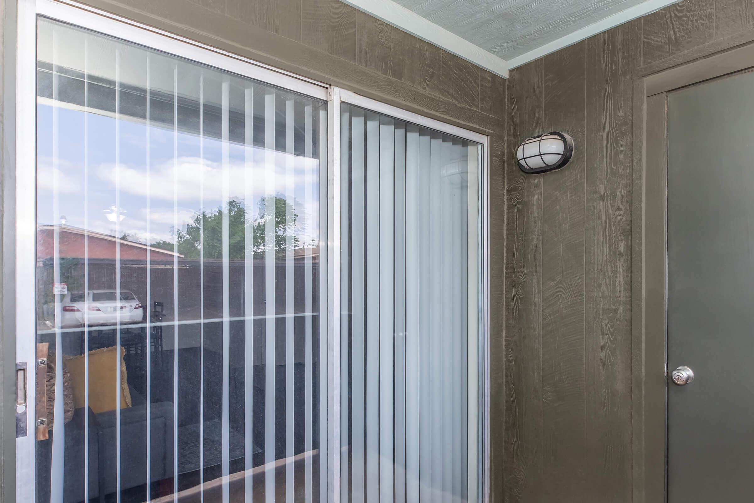 Sliding glass door with vertical blinds, showing reflections of the outside. The walls are painted a dark color, and a light fixture is mounted on one side. The image captures a corner of an outdoor patio area, partially visible through the door.