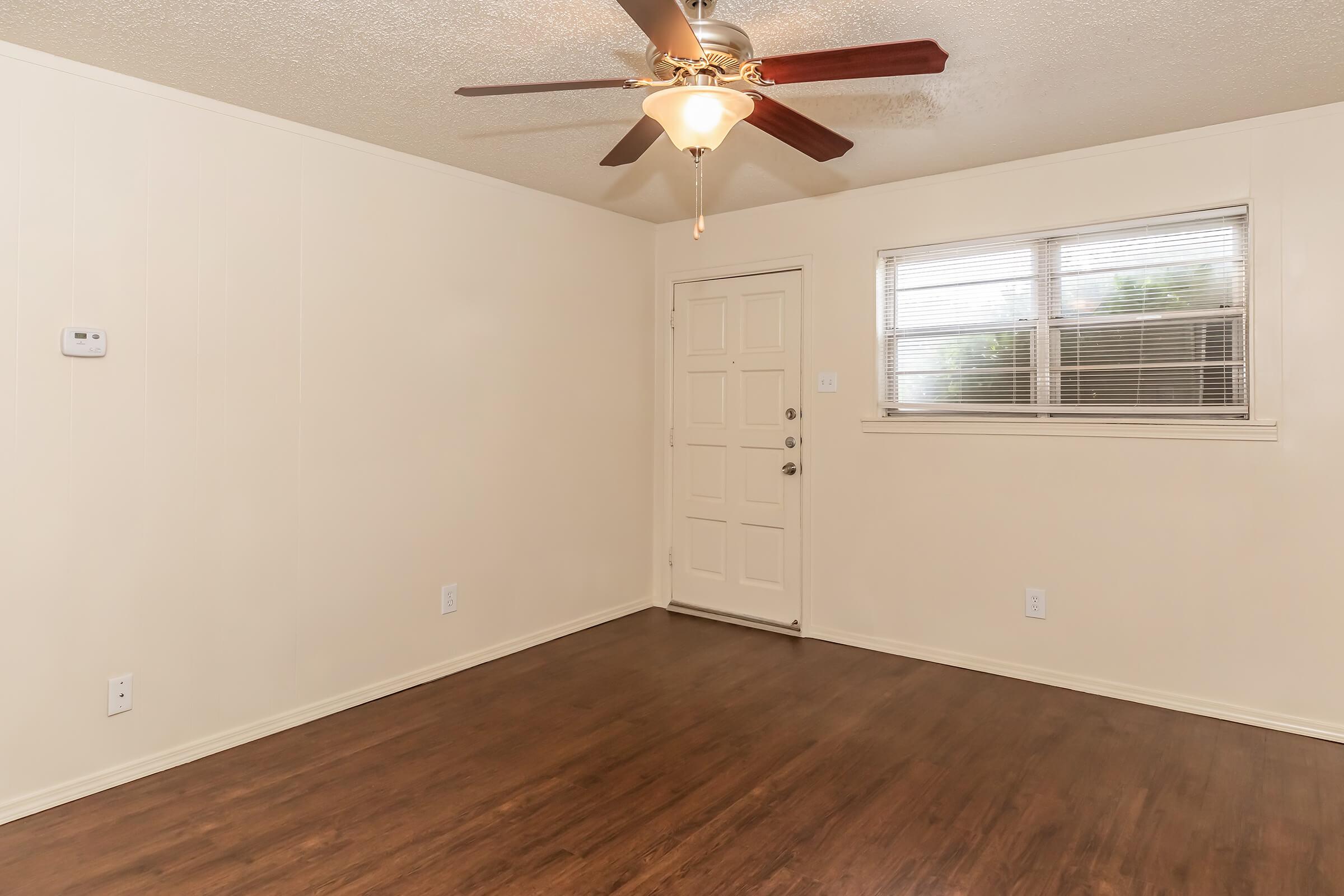 A spacious, empty room with light-colored walls and a brown wood-like floor. A ceiling fan hangs from the ceiling, and there’s a window on one side. The front door is visible, featuring a simple design with a small window pane. Overall, the room appears bright and ready for furnishing.
