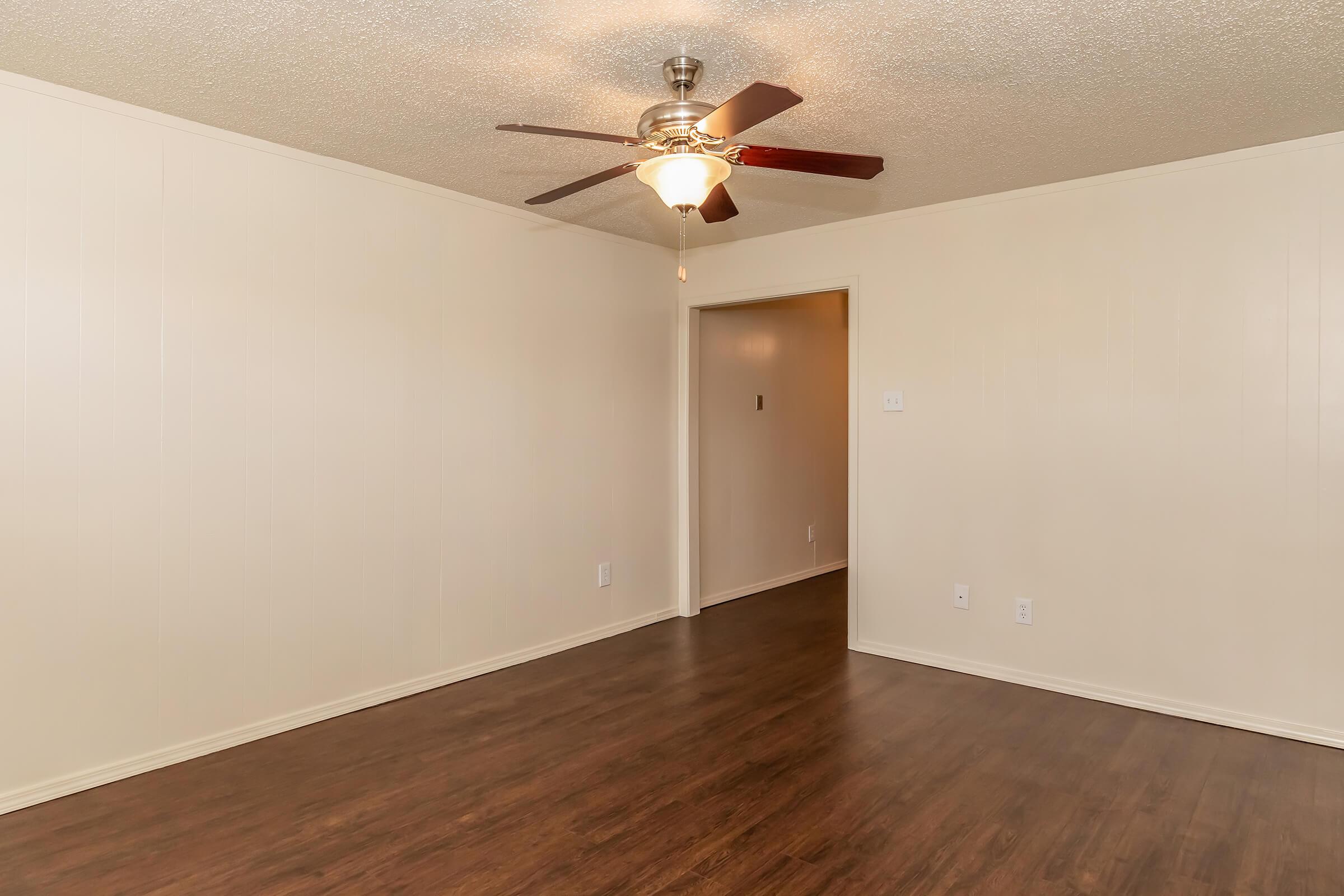 A spacious, empty room with light-colored walls and dark wooden flooring. The ceiling features a ceiling fan with light. A doorway leads to another room visible in the background, and the overall ambiance is bright and airy.