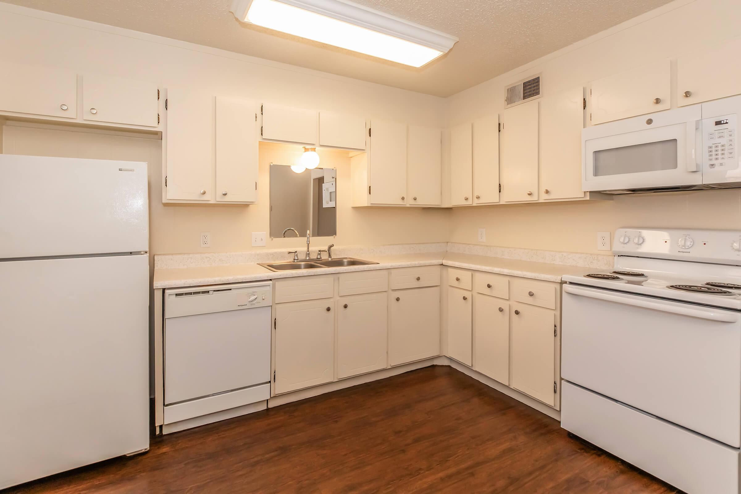 A well-lit kitchen featuring white cabinetry, a stainless steel sink, and modern appliances including a refrigerator, dishwasher, microwave, and stove. The floor has a dark wood finish, and there is a mirror above the sink, creating a clean and inviting space.