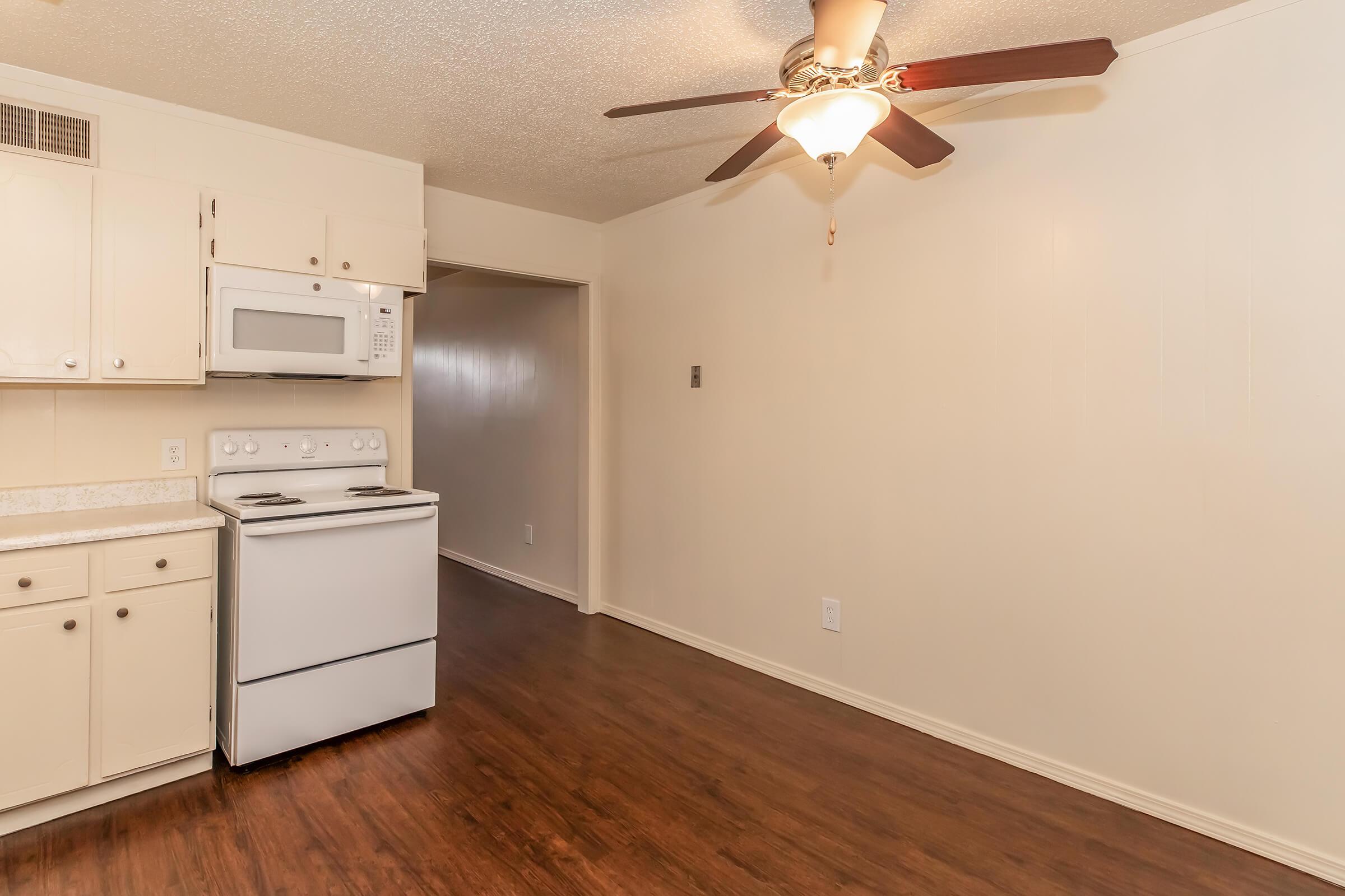 A small kitchen area featuring white cabinets, a white stove, and a built-in microwave. The kitchen has a ceiling fan and is adjacent to a hallway. The floor is dark wood, and the walls are painted a light color, creating a simple and clean appearance.