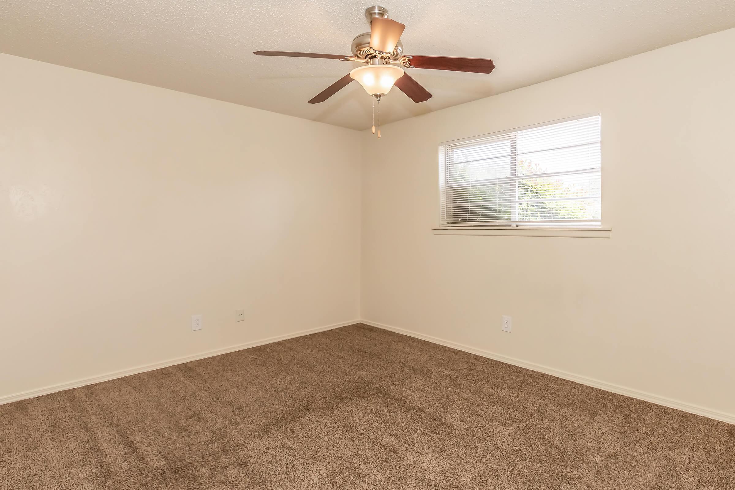 A spacious, unfurnished room featuring light beige walls and a ceiling fan with wooden blades. The carpeted floor is a similar shade, and there is a window with blinds allowing natural light to enter. The room appears clean and well-maintained, ideal for various uses.