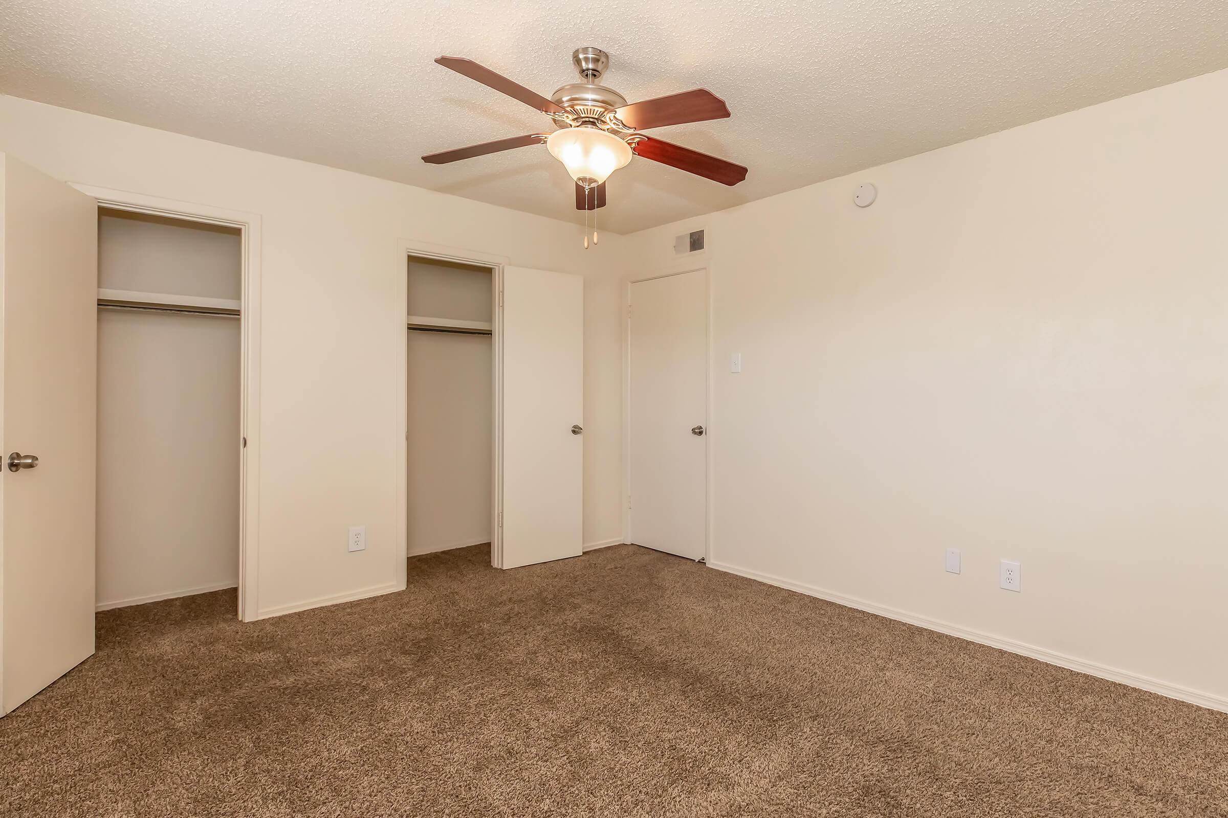 A view of a neutral-colored bedroom featuring beige walls and carpet. The room has a ceiling fan with wooden blades, two closet doors on the left, and a closed door on the right. Natural light illuminates the space, creating a warm and inviting atmosphere.