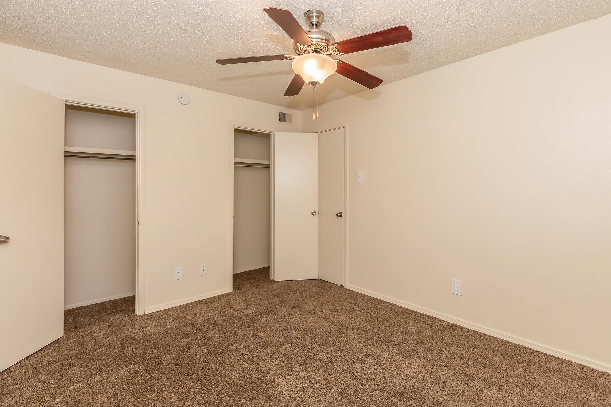 A well-lit bedroom featuring beige walls and carpet, with a ceiling fan. There are two closed doors leading to closets, and the room appears empty and spacious, creating a cozy atmosphere.