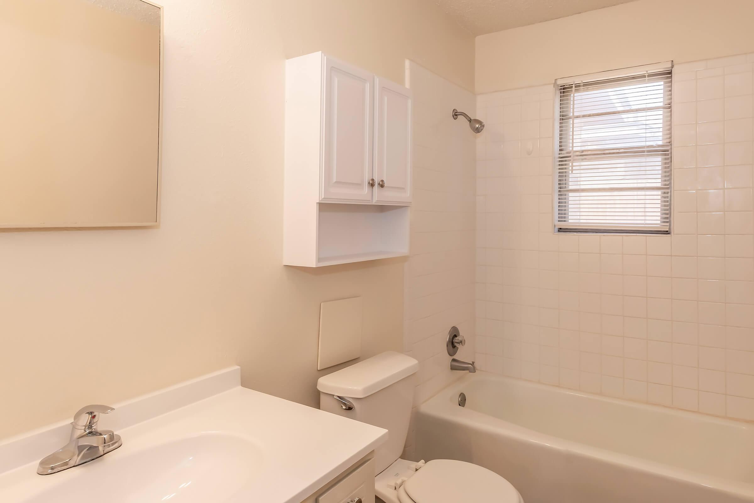 A clean, modest bathroom featuring a white bathtub, shower, and toilet. A small white cabinet is mounted above the sink, which has a single faucet and a rectangular mirror above it. Natural light enters through a window with blinds, illuminating the tiled walls.