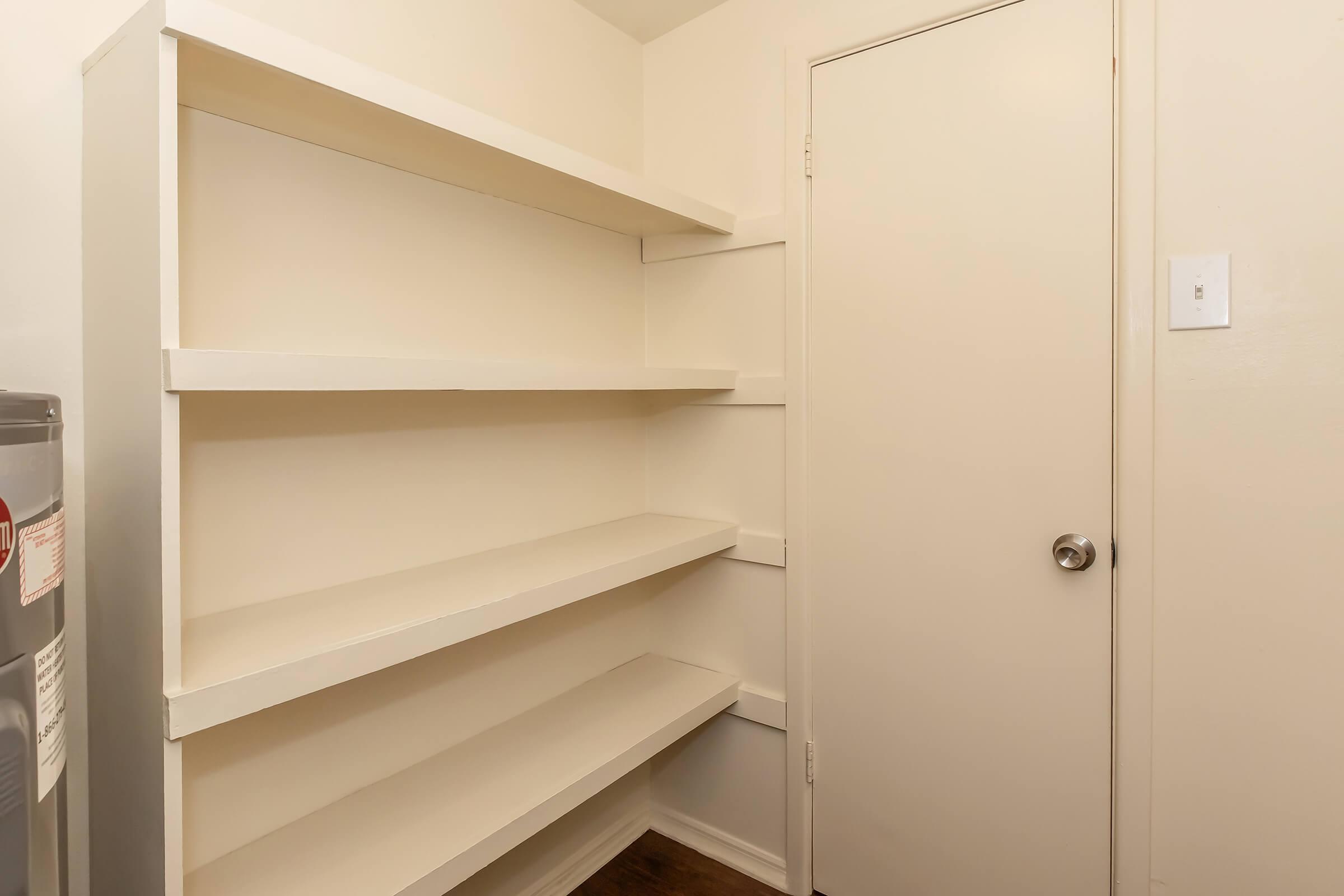 Empty room with a white wall-mounted shelving unit on the left side, featuring three horizontal shelves. A closed beige door is visible on the right side. The flooring is wooden. The space appears to be tidy and uncluttered, suggesting it may be used for storage.
