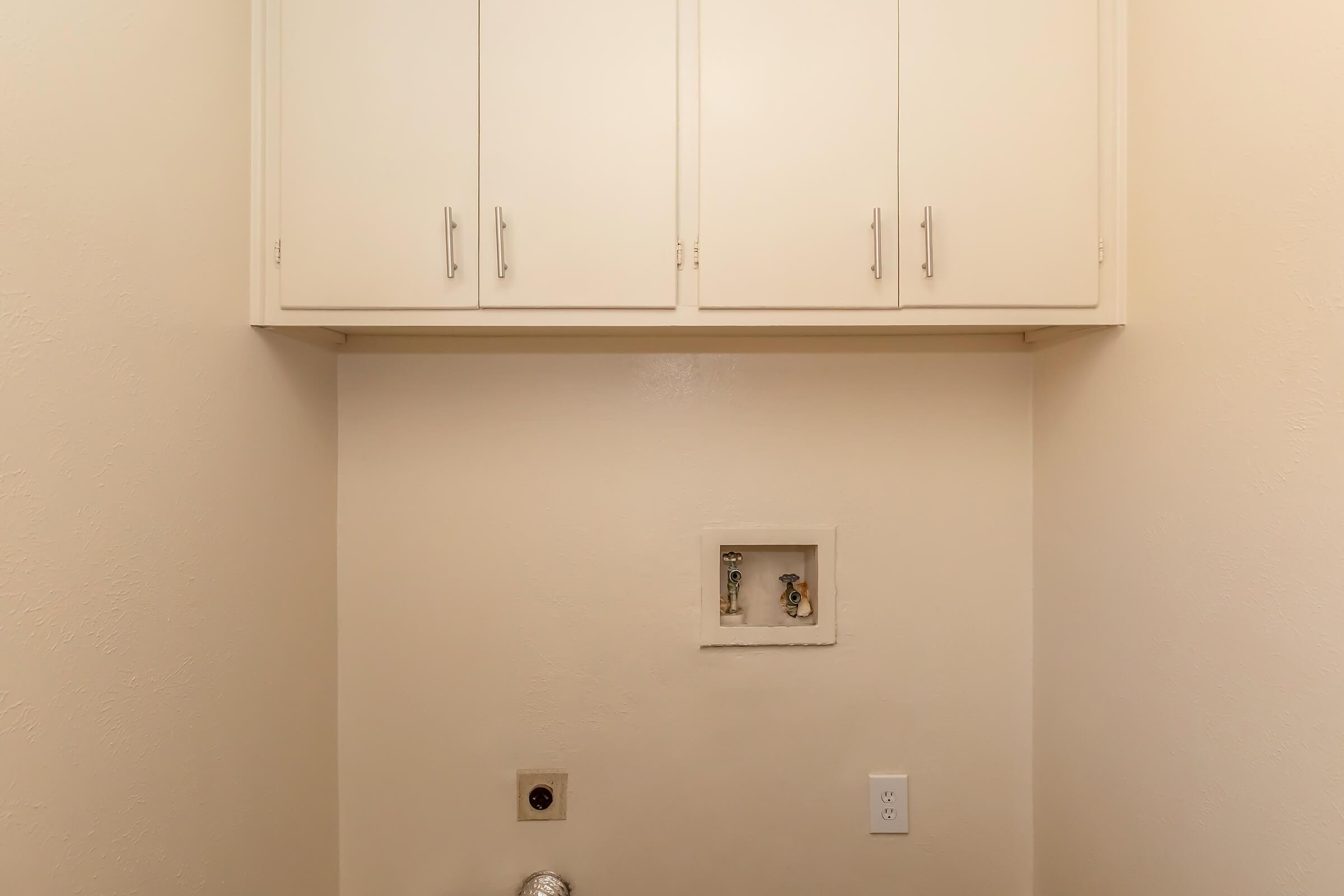 A clean, light-colored laundry room featuring white cabinets above, with a space for a washing machine and dryer below. There is an electrical outlet and a small panel for hookups on the wall, providing a minimalist and functional design. The walls are a soft, neutral tone.