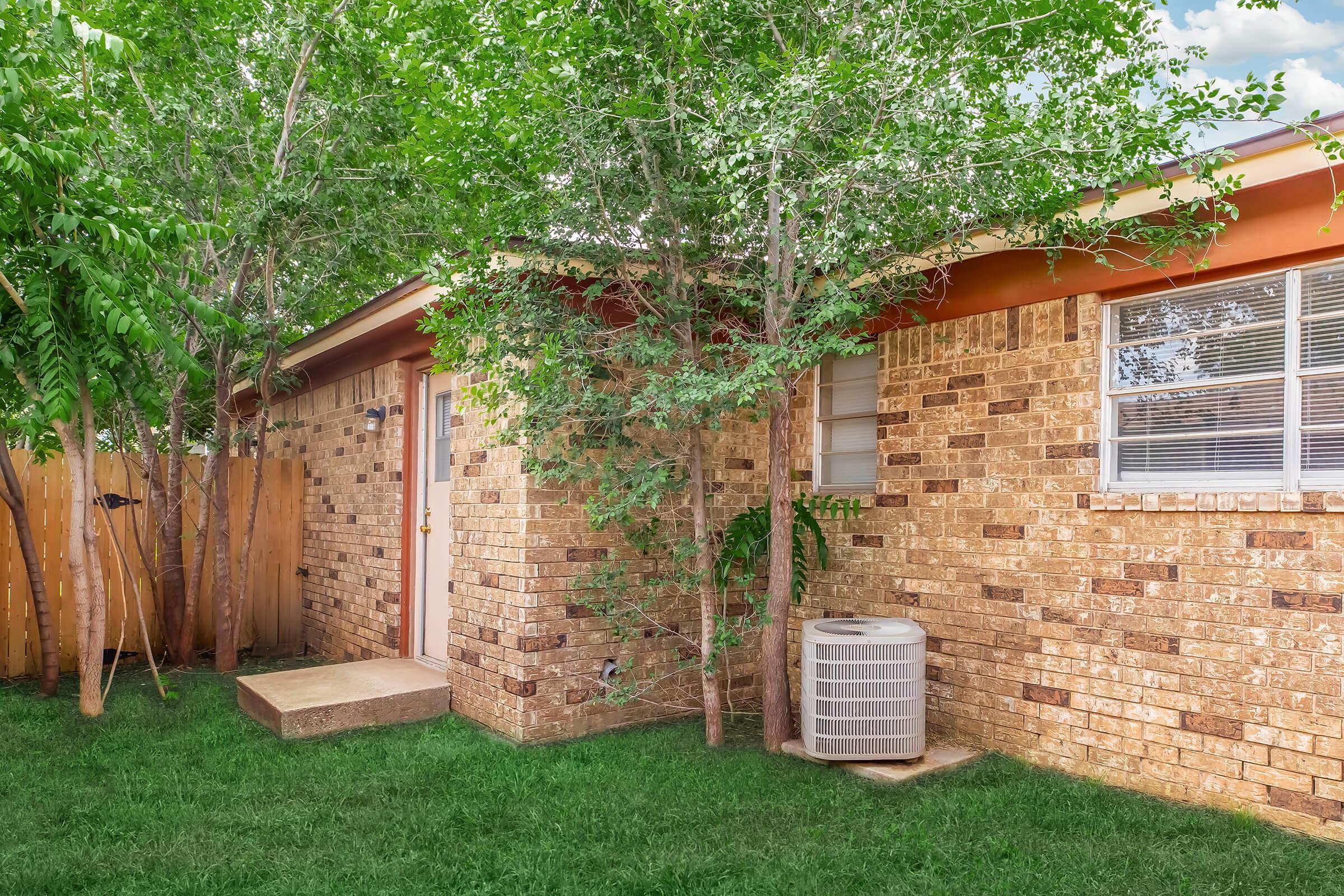 A brick exterior of a house with a small porch, surrounded by green grass and trees. There is an air conditioning unit on the ground beside the house, and a wooden fence in the background partially obscures the view. The sky is bright with scattered clouds.